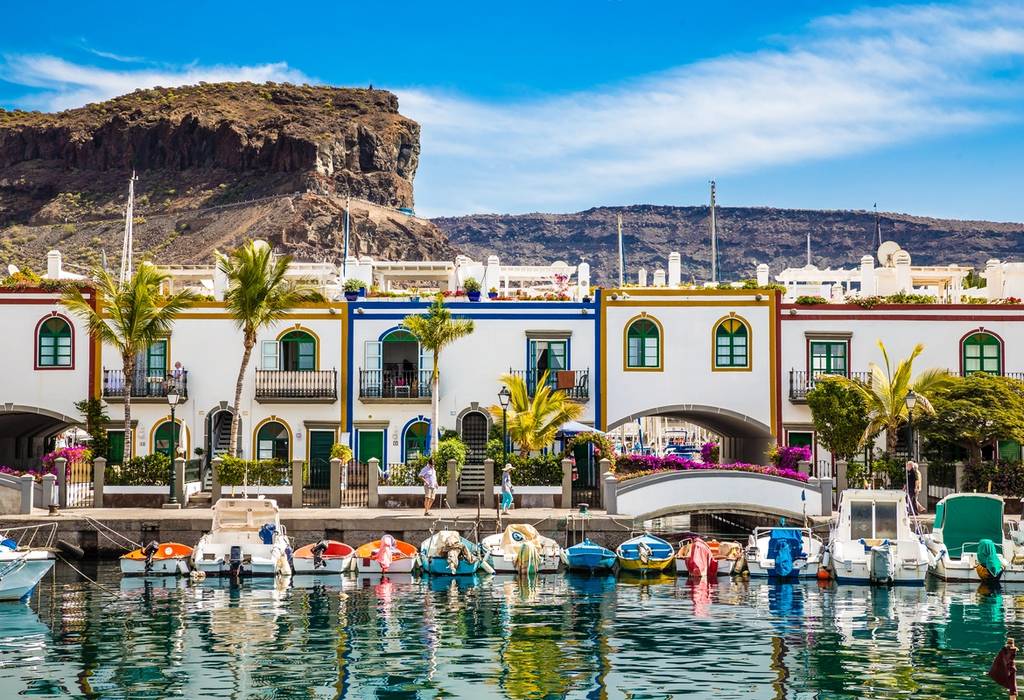 The traditional white buildings with colourful trim in Puerto de Mogan, with boats moored in the front and a backdrop of mountains