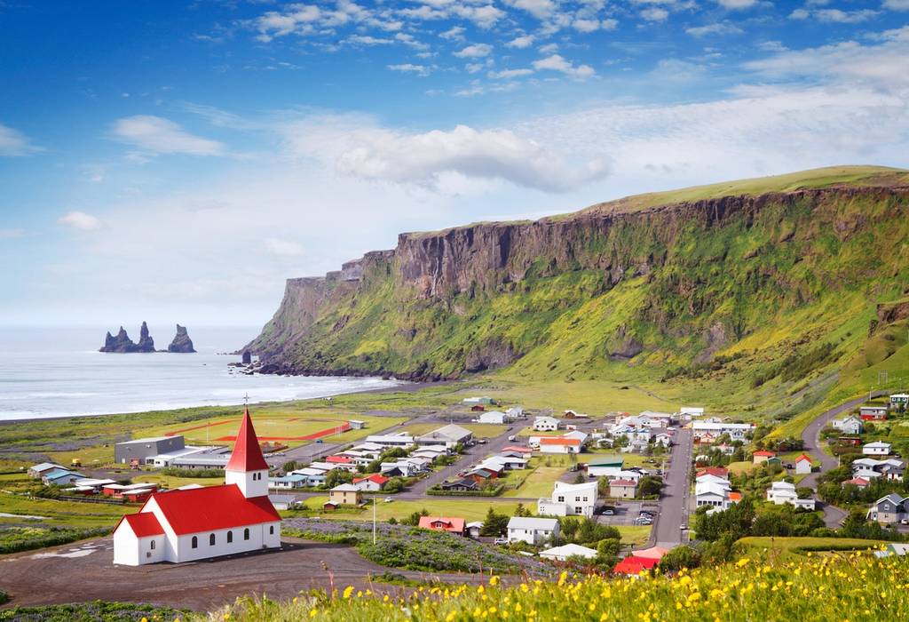 View of a white and red church overlooking a small town at the foot of dramatic cliffs with the sea stacks of Vik beach in background