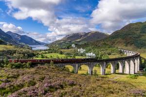 View of the famous red Jacobite Steam Train over the curved Glenfinnan Railway Viaduct in Scotland with a loch in the background and green hills on all sides