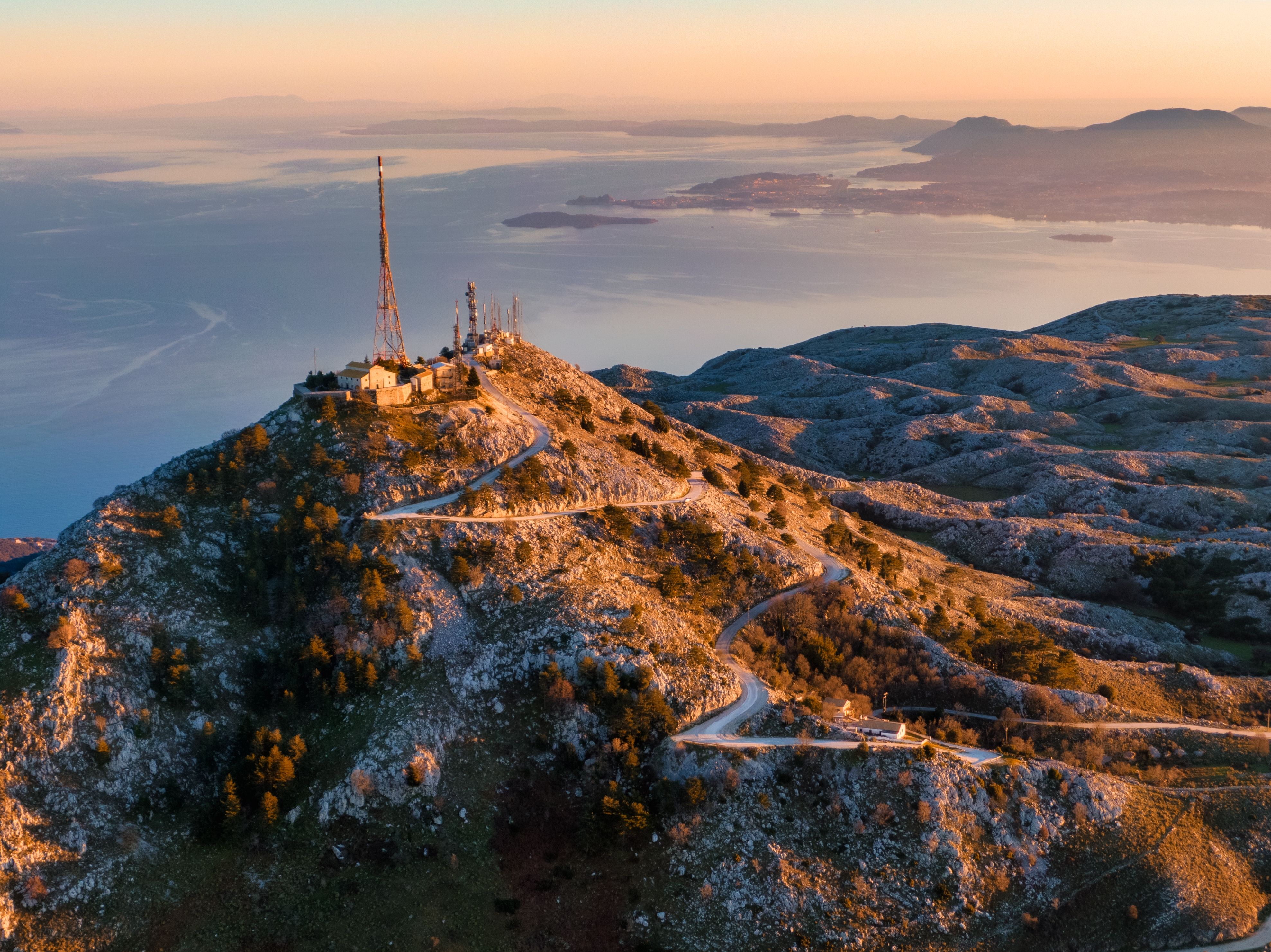 Sunset drone view of a winding road leading up towards a telecommunications station with Corfu town in background.