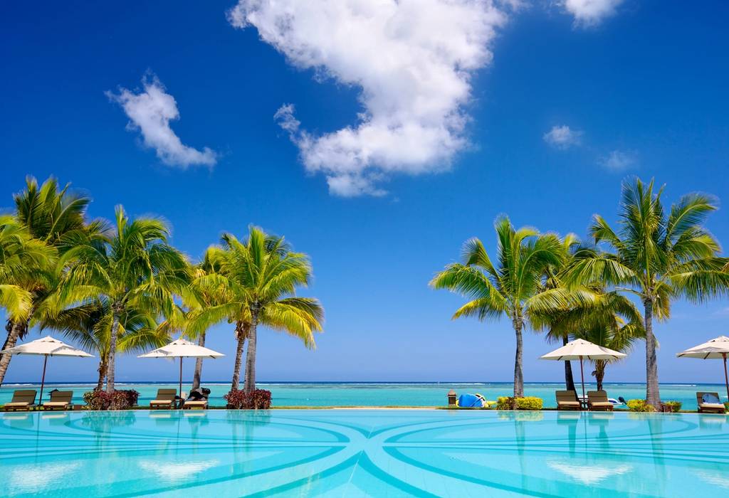 A view across a resort swimming pool looking out to sea in Mauritius with sun loungers, umbrellas and palm trees