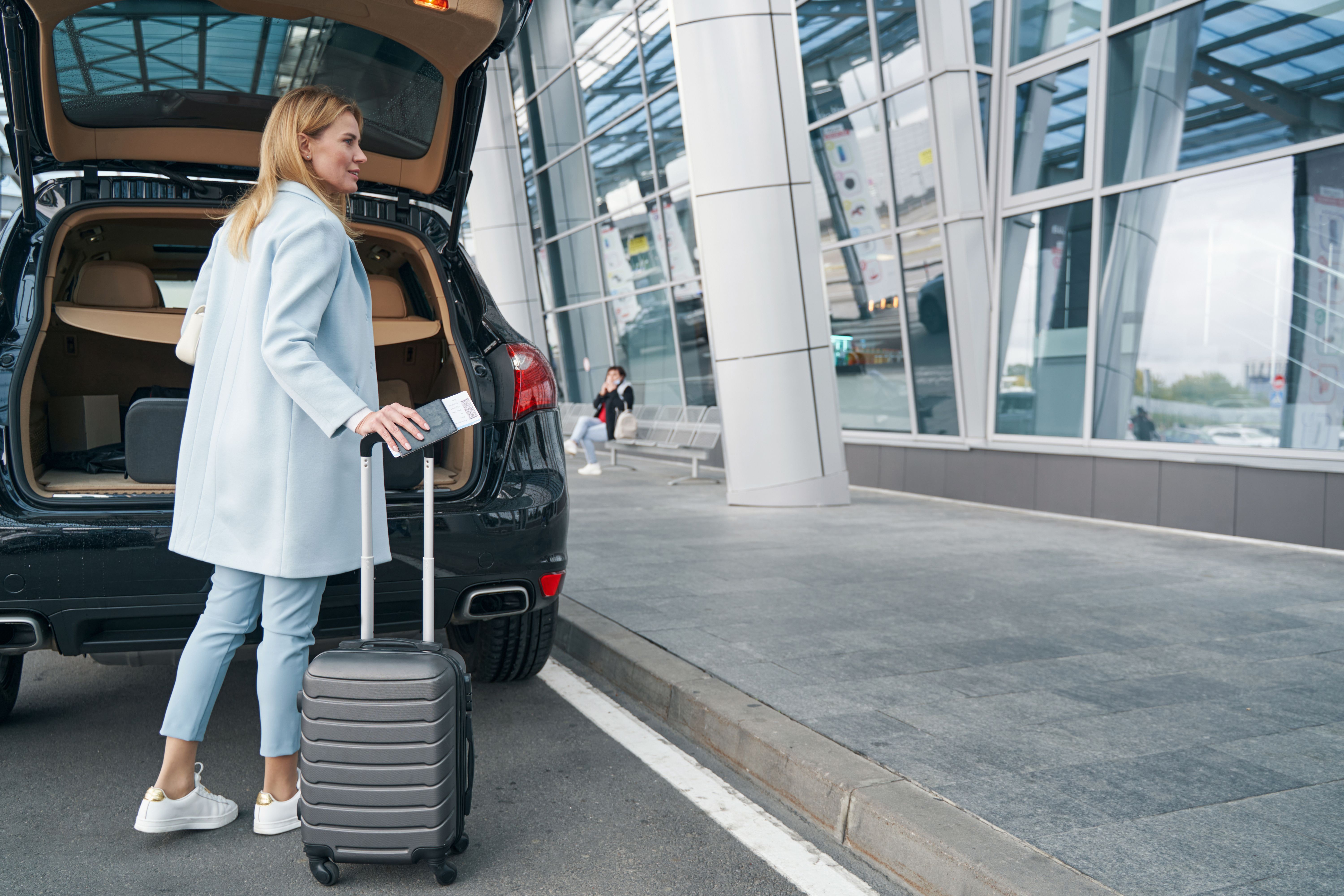 A woman stands at the back of a vehicle holding a boarding ticket and a trolley suitcase.