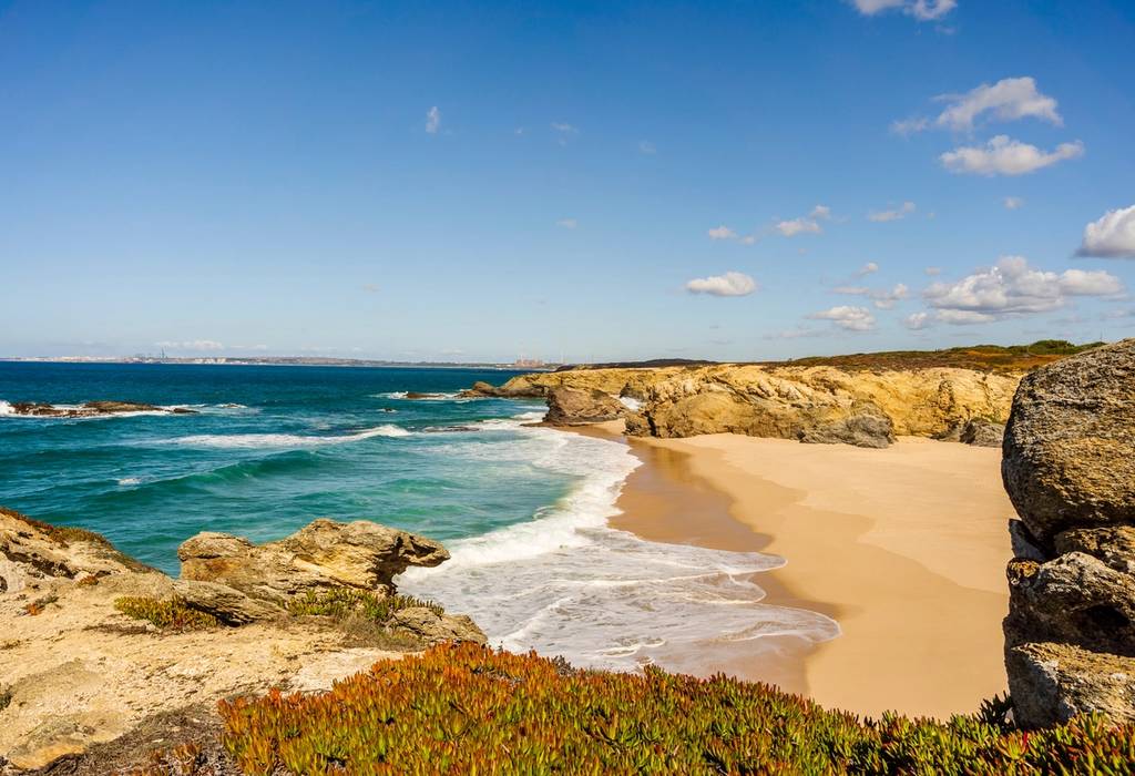 An empty, sandy beach hemmed in by rocky cliffs on the west coast of Portugal on a sunny day