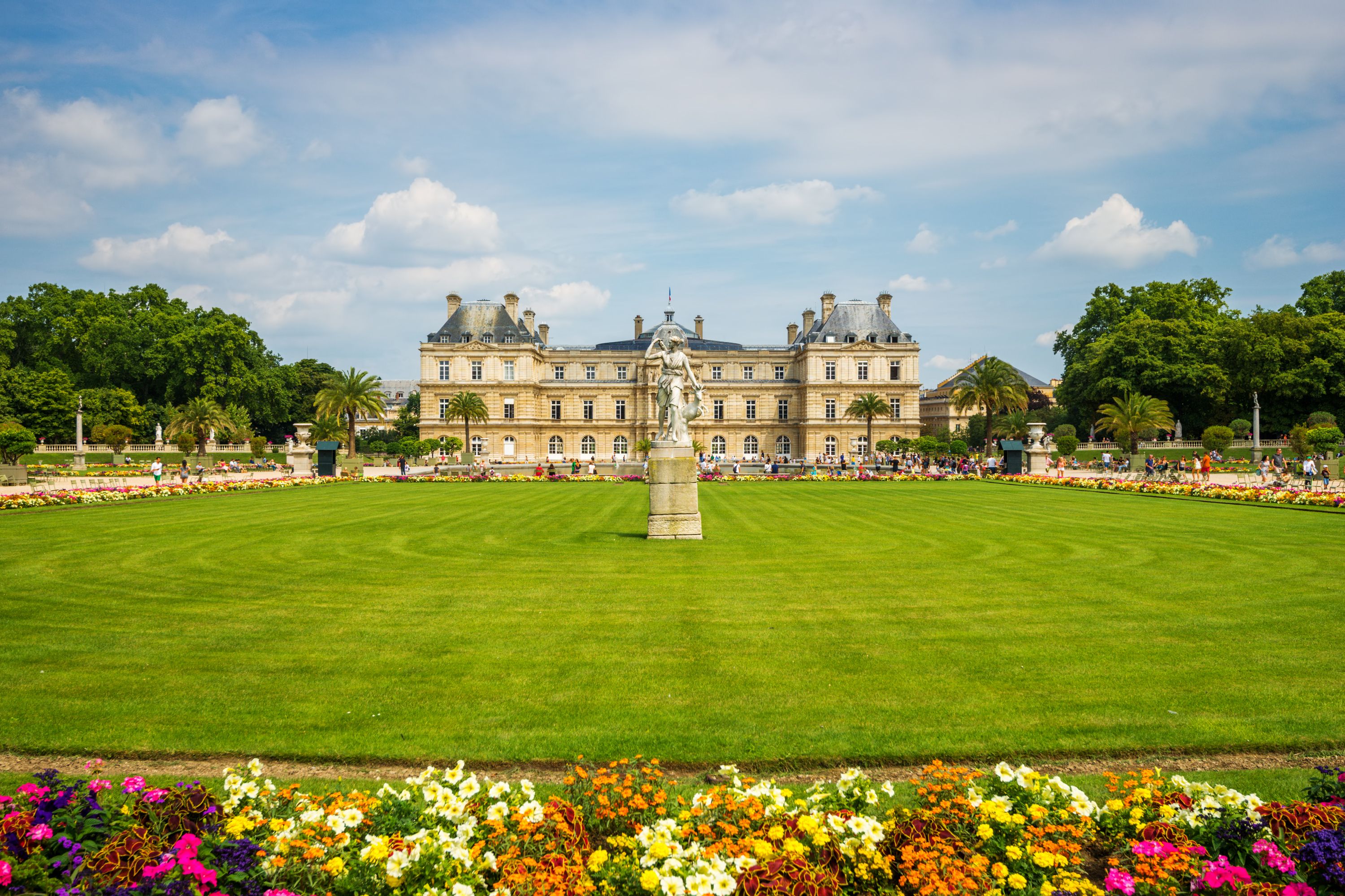 A view of Luxembourg gardens and Palace in Paris with colourful flowers and a lush green lawn with a statue in the centre