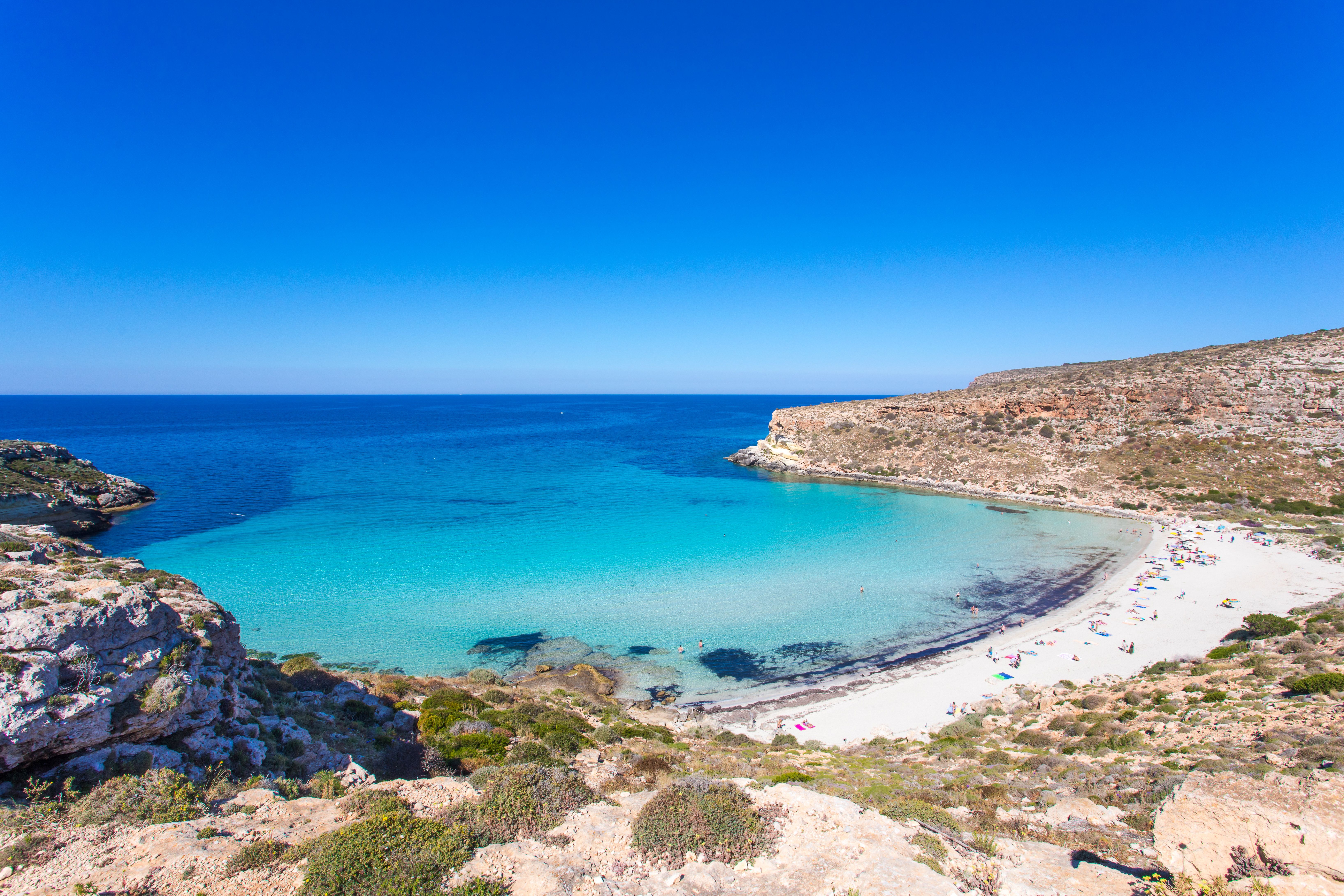Aerial view of a sandy, sun-baked beach lapped by brilliant blue water