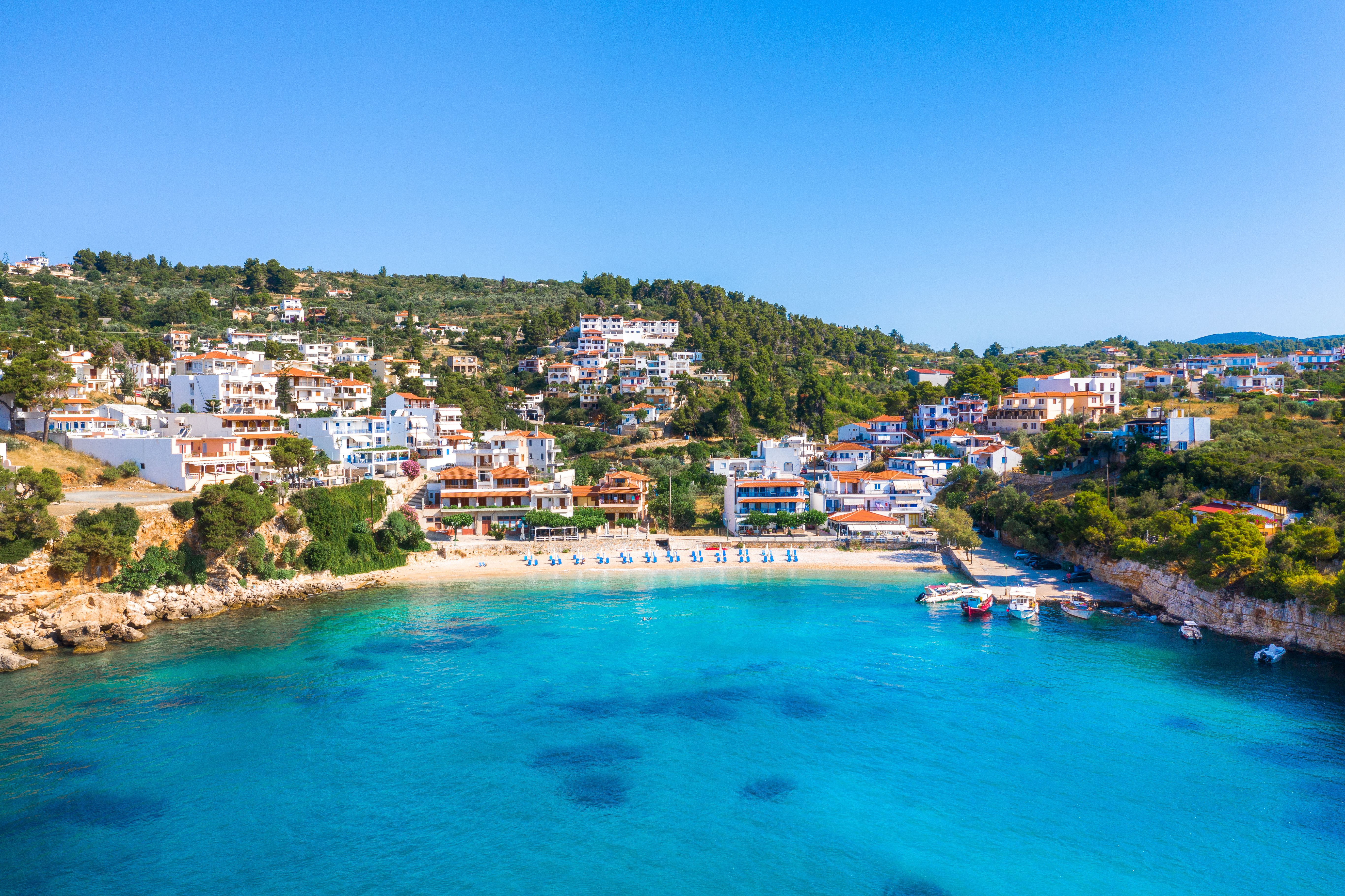 Drone view from the sea looking towards a sun-lounger-lined golden beach backed by a small whitewashed town.
