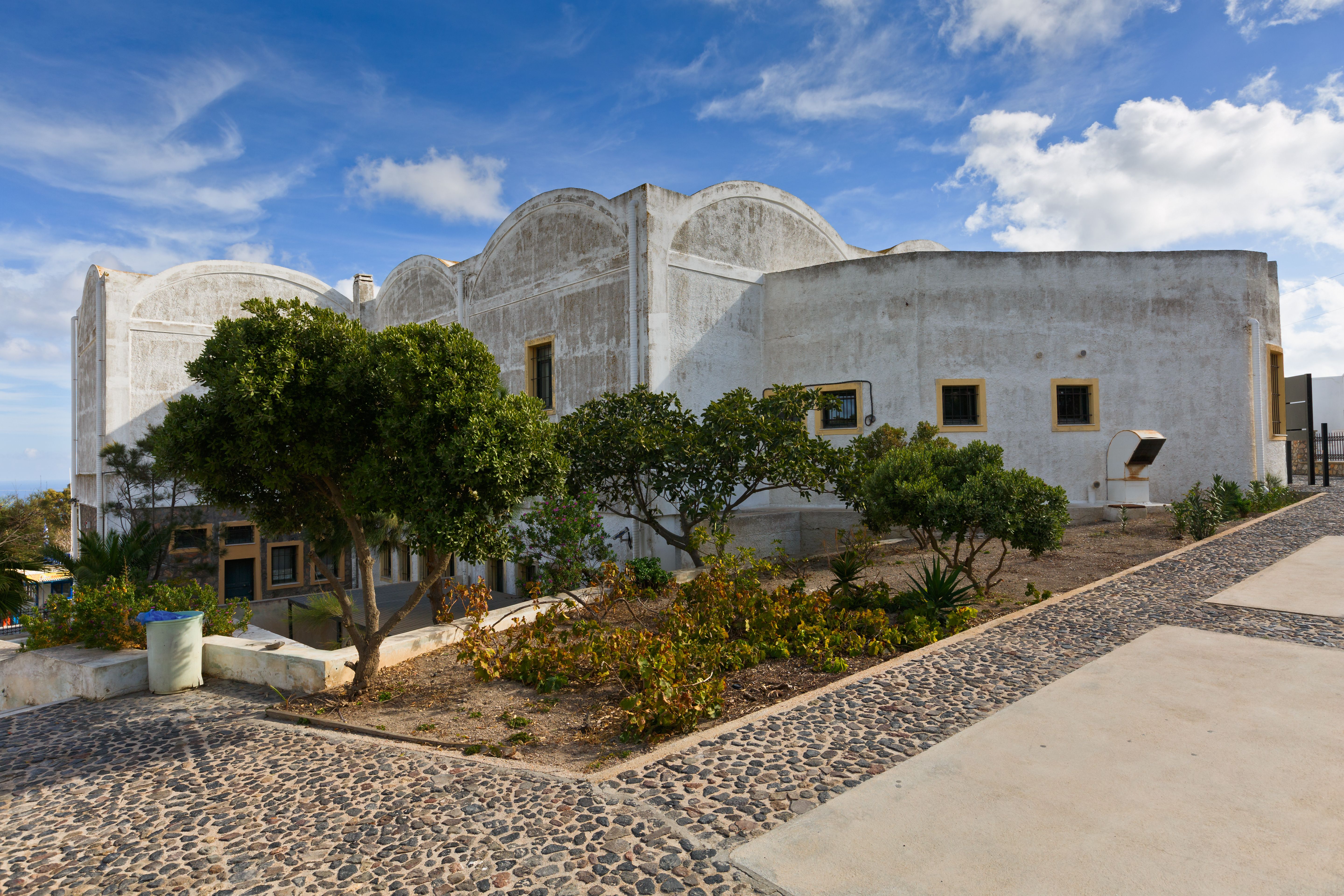 A grey stone building with a small garden of trees in the front on a sunny day