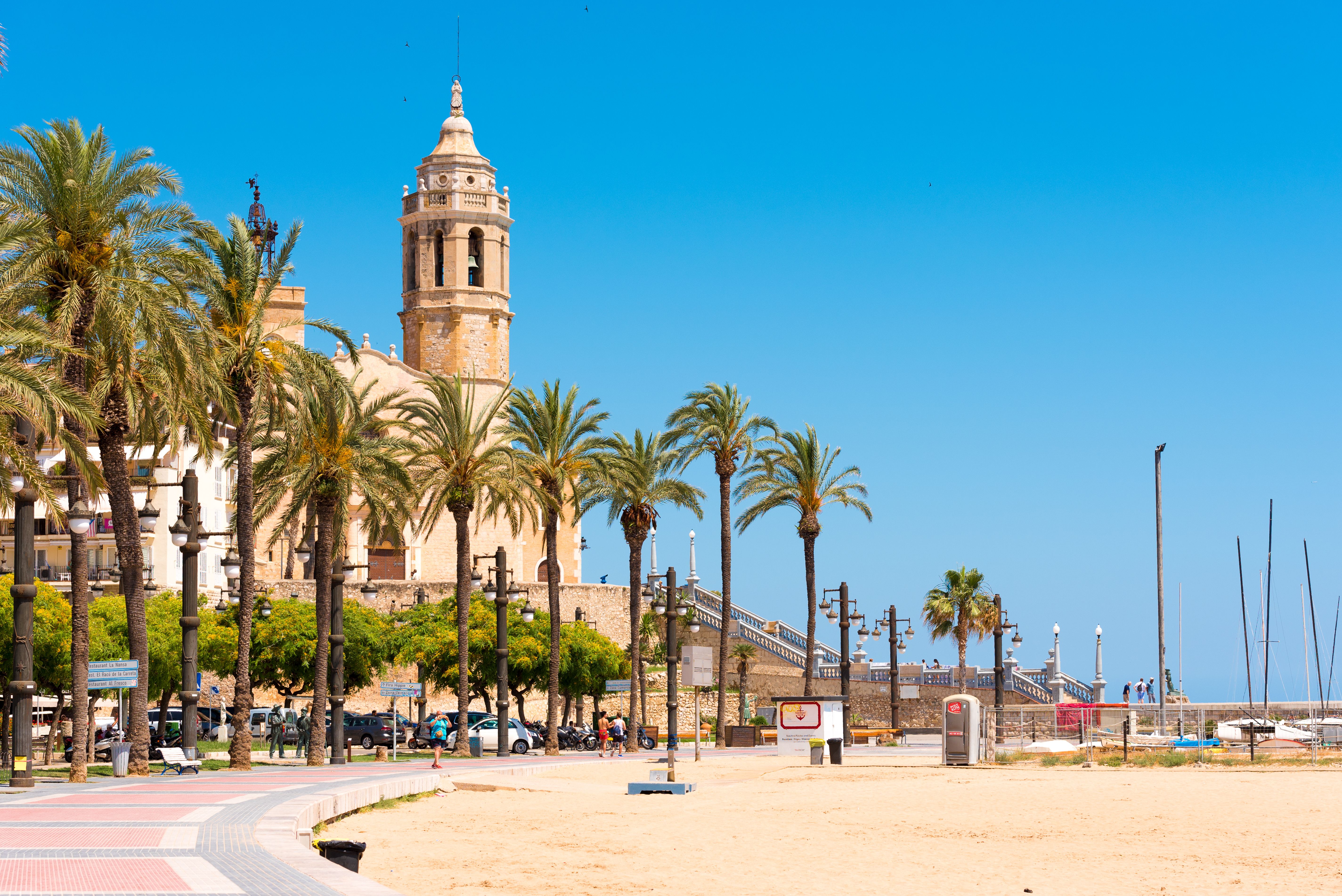 View of golden sands and a curved promenade lined with palm trees with a honey-coloured church in the background