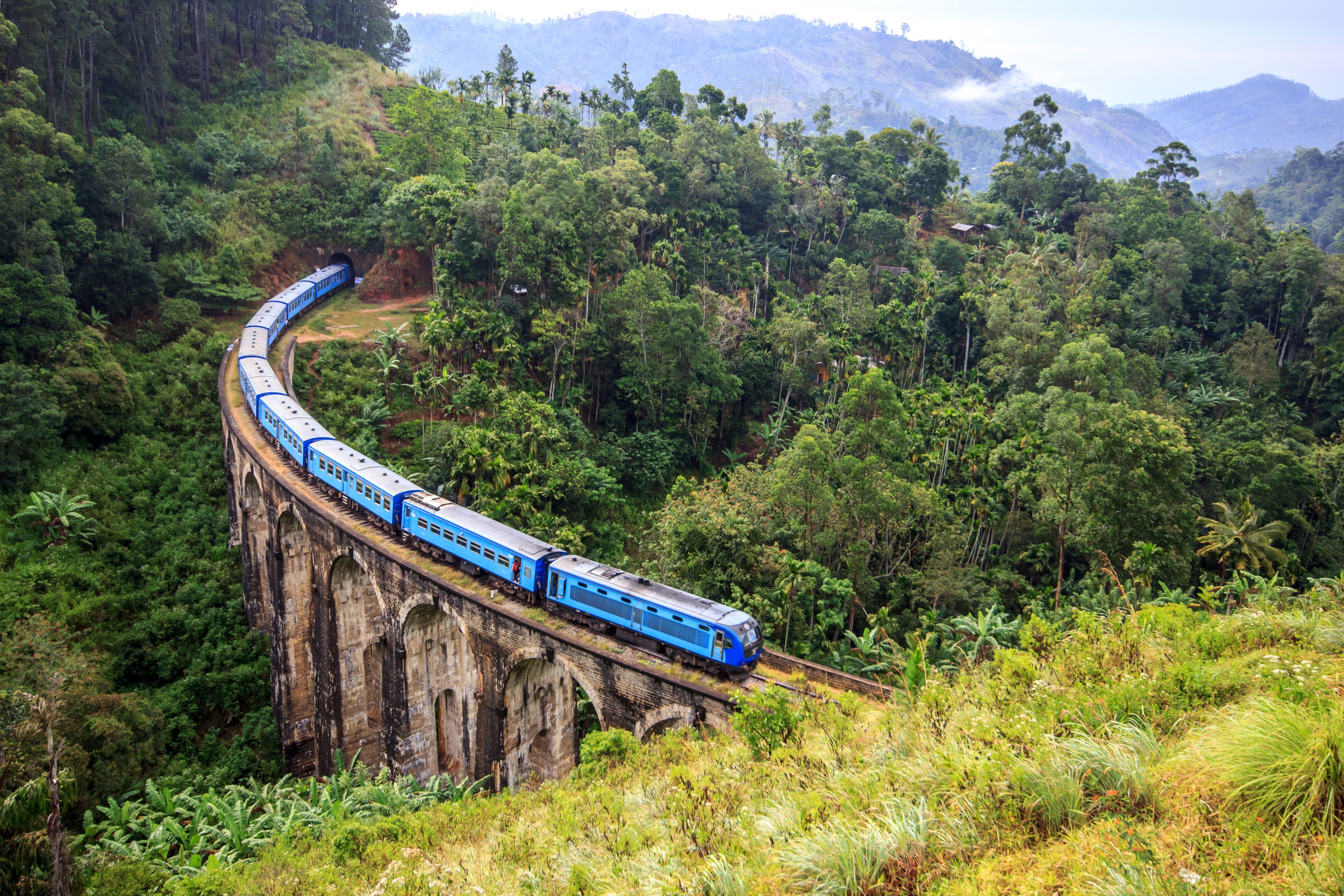 View of a blue train travelling across a huge arched bridge in a forested area in Sri Lanka