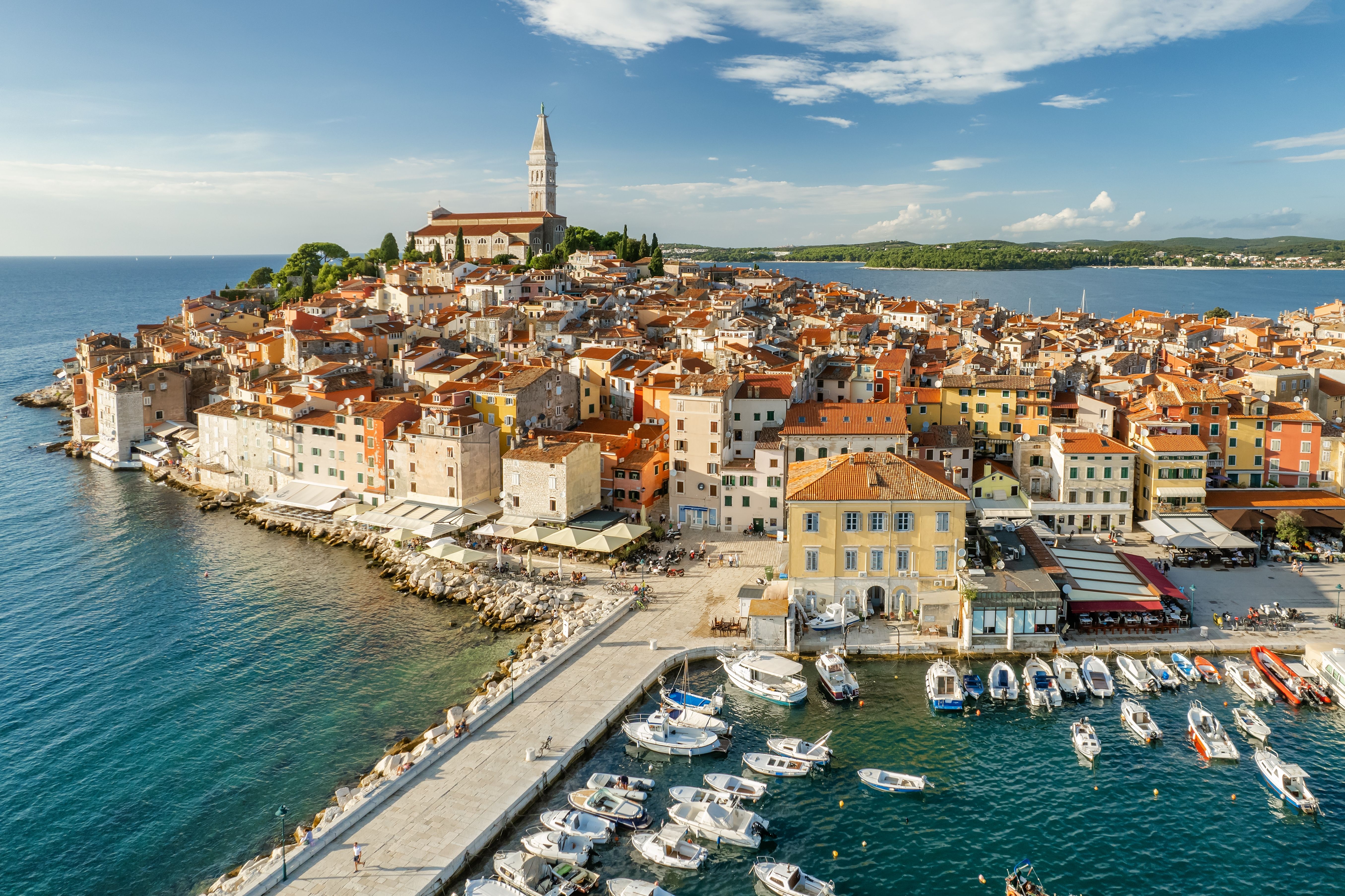 Aerial view of a a white and pastel-yellow old town perched on a little promontory with a cathedral at its highest point.