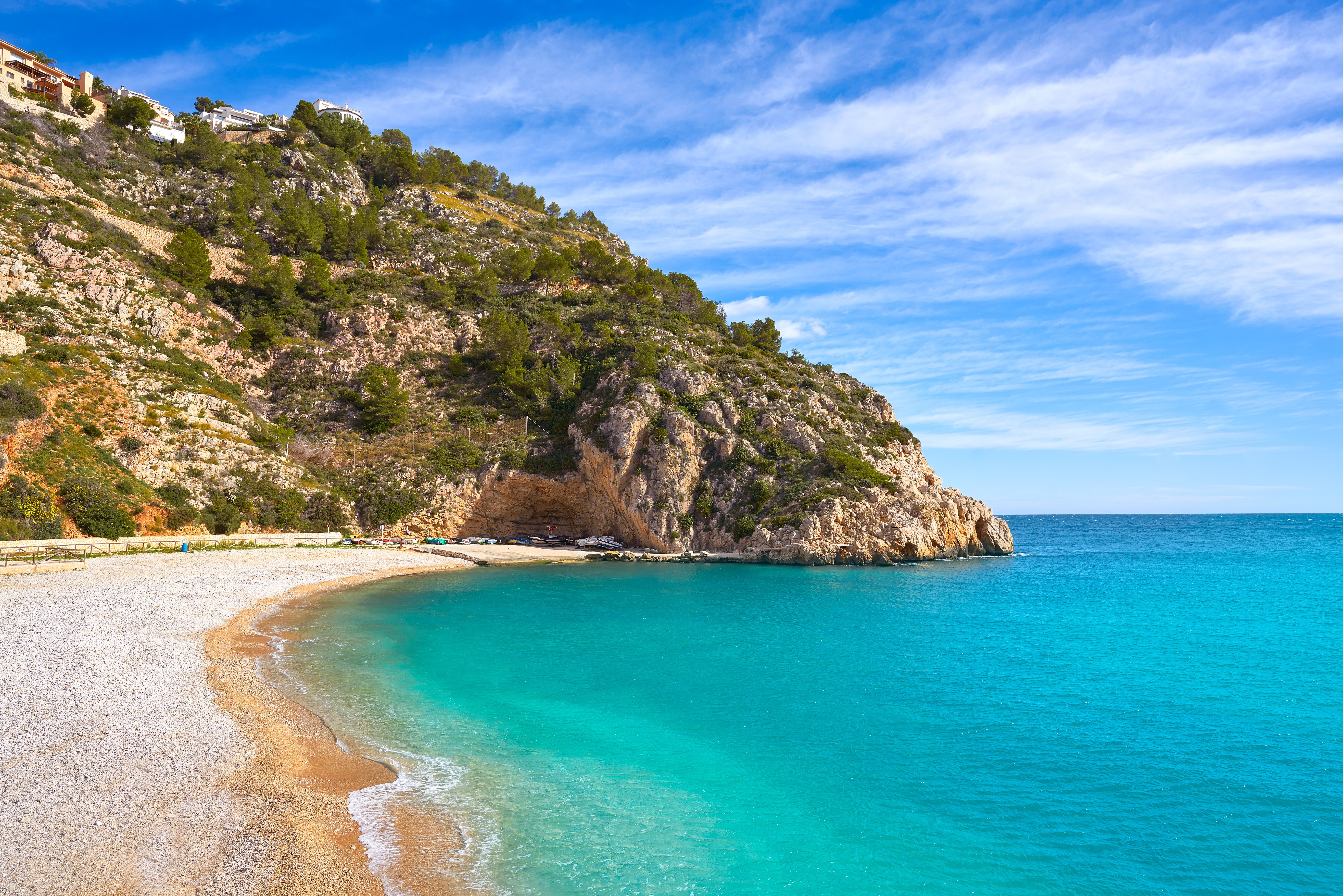 View of an empty sand-and-pebble beach with a towering tree-covered cliff on the far end.