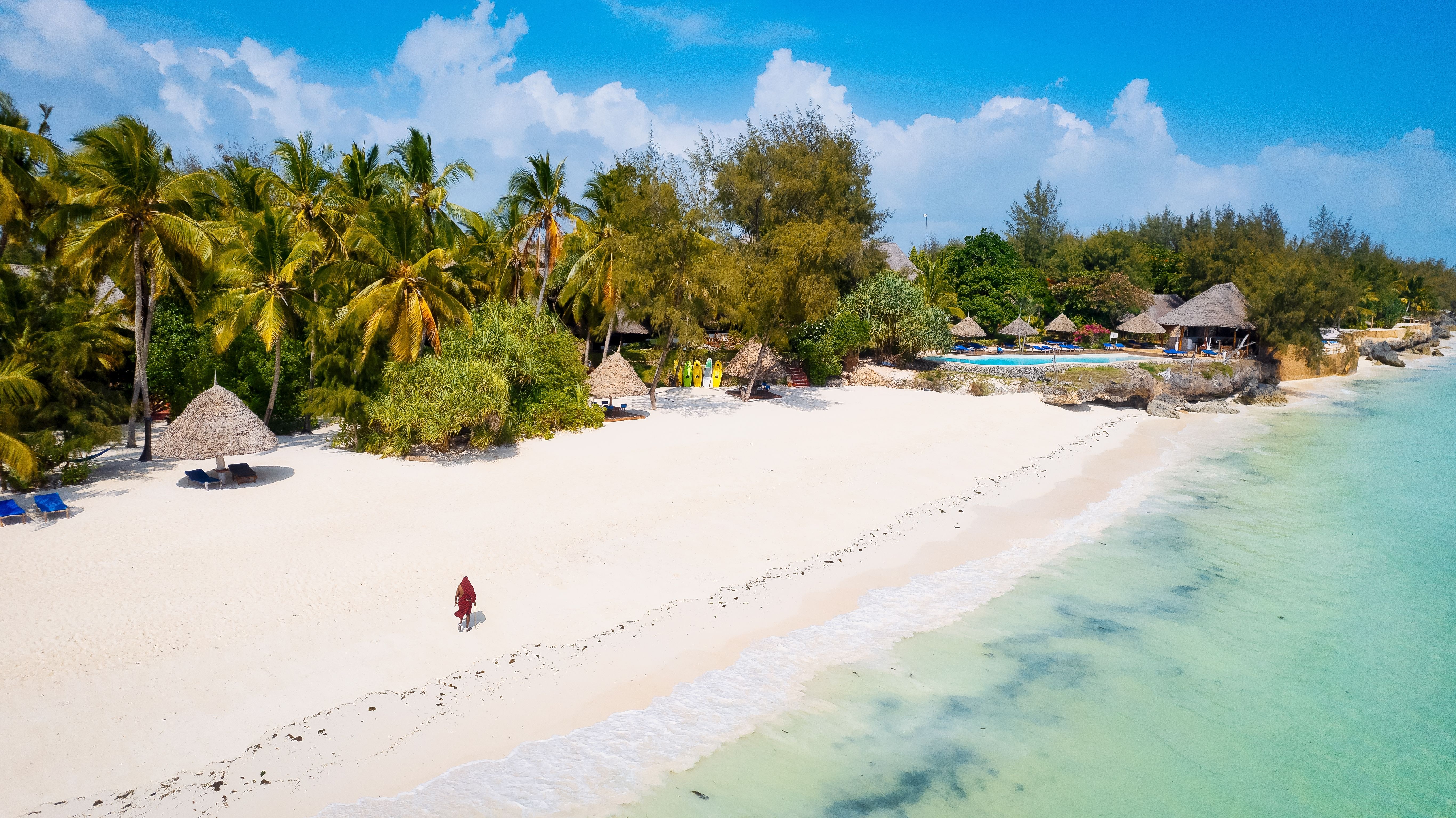 A person walks along a pristine white-sand beach backed by swaying palms and lapped by baby blue waters