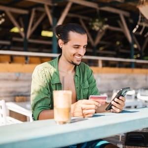 A picture of a man at a beach bar with a coffee, whilst holding a card to pay, and looking at his phone smiling