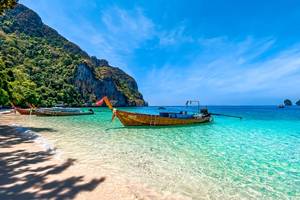 Wooden long-tail boats wait in the clear waters and on the golden sands of Monkey Beach in Koh Phi Phi Don