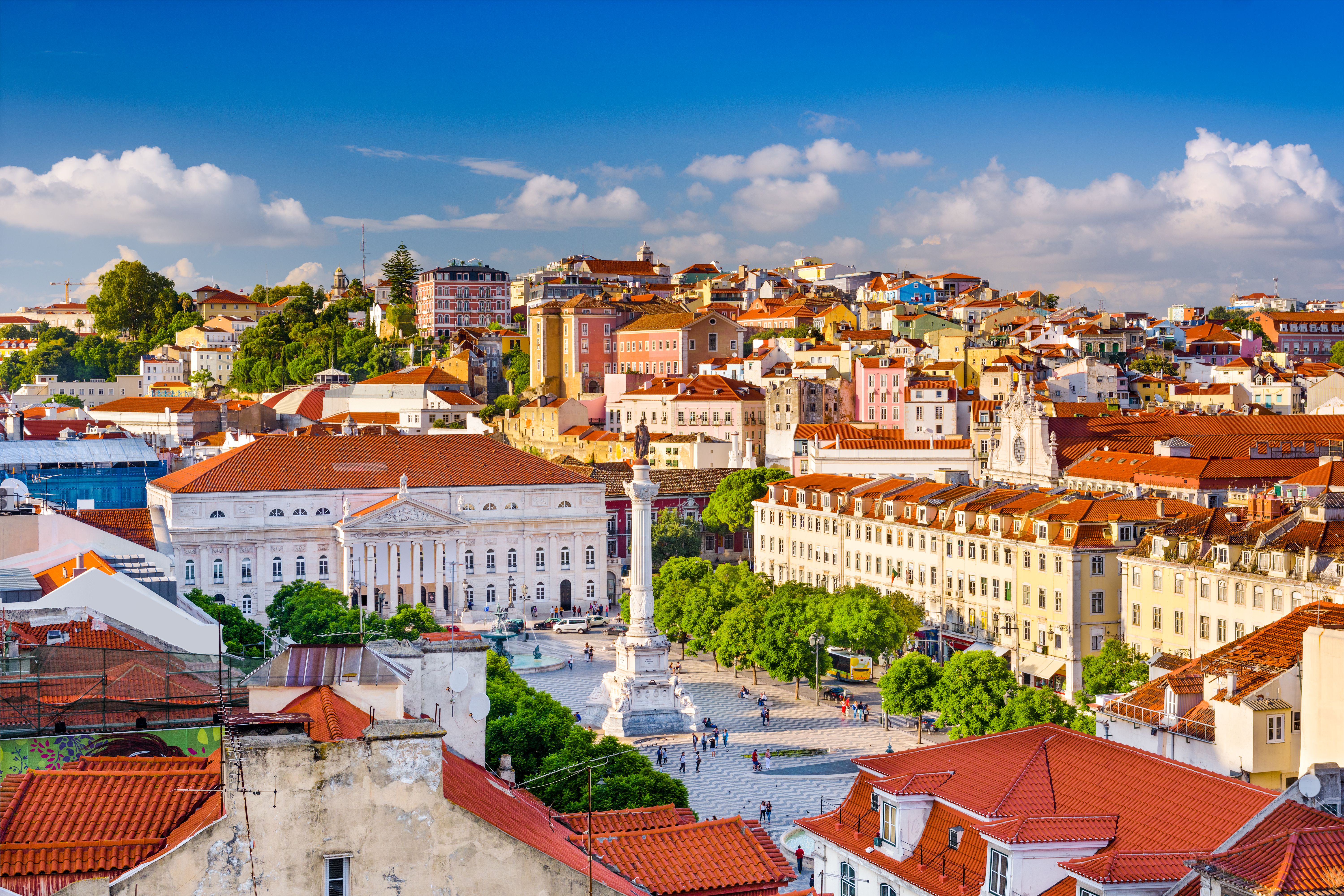 Aerial view of a square in Lisbon with a tall columned monument in the centre and trees and grand buildings surrounding it.