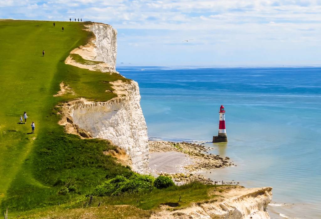 The grass-covered white chalk cliffs of the South Downs with a red and white striped lighthouse below in the English Channel