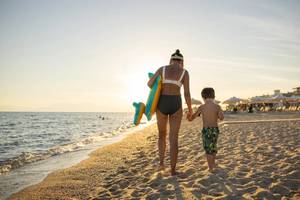 Mother and child walking on a beach with the mother holding an inflatable toy and deck chairs and umbrellas in the distance