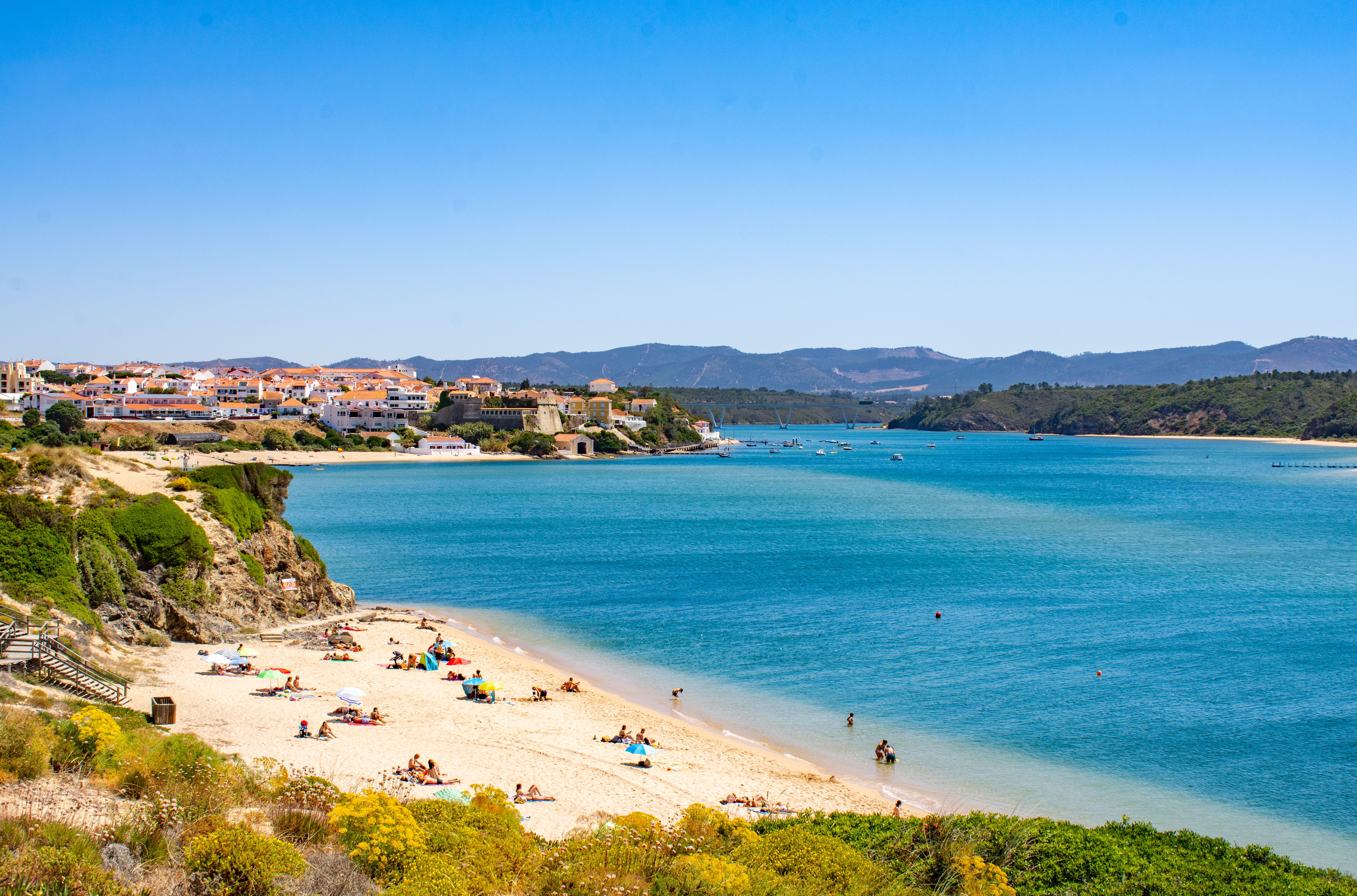 View of sunbathers on a small, sandy beach on the mouth of a river with a town in the background.