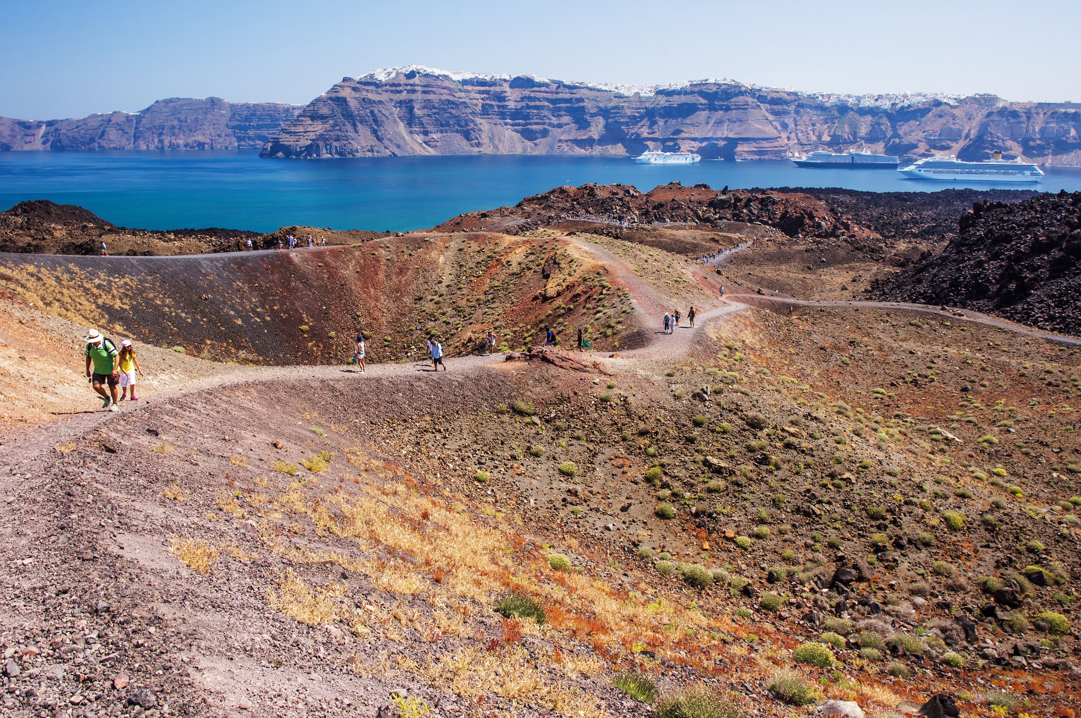Hikers walking along the narrow dirt paths across a volcanic landscape in Santorini with cruise ships moored in the sea in the background