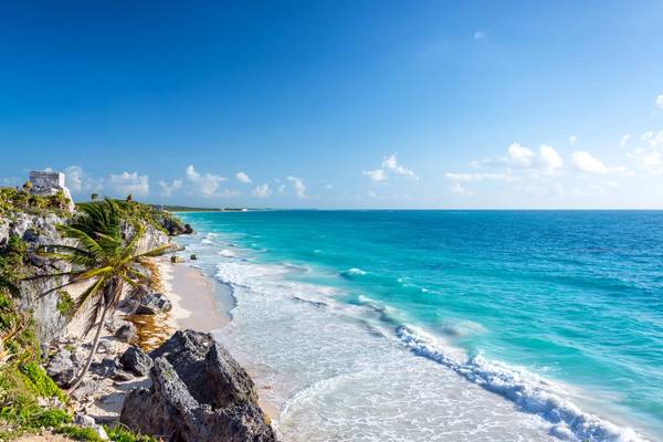 Wide-angle view of the frothy turquoise waters of the Caribbean Sea, a small white-sand beach with a palm tree and the ruins of Tulum, Mexico