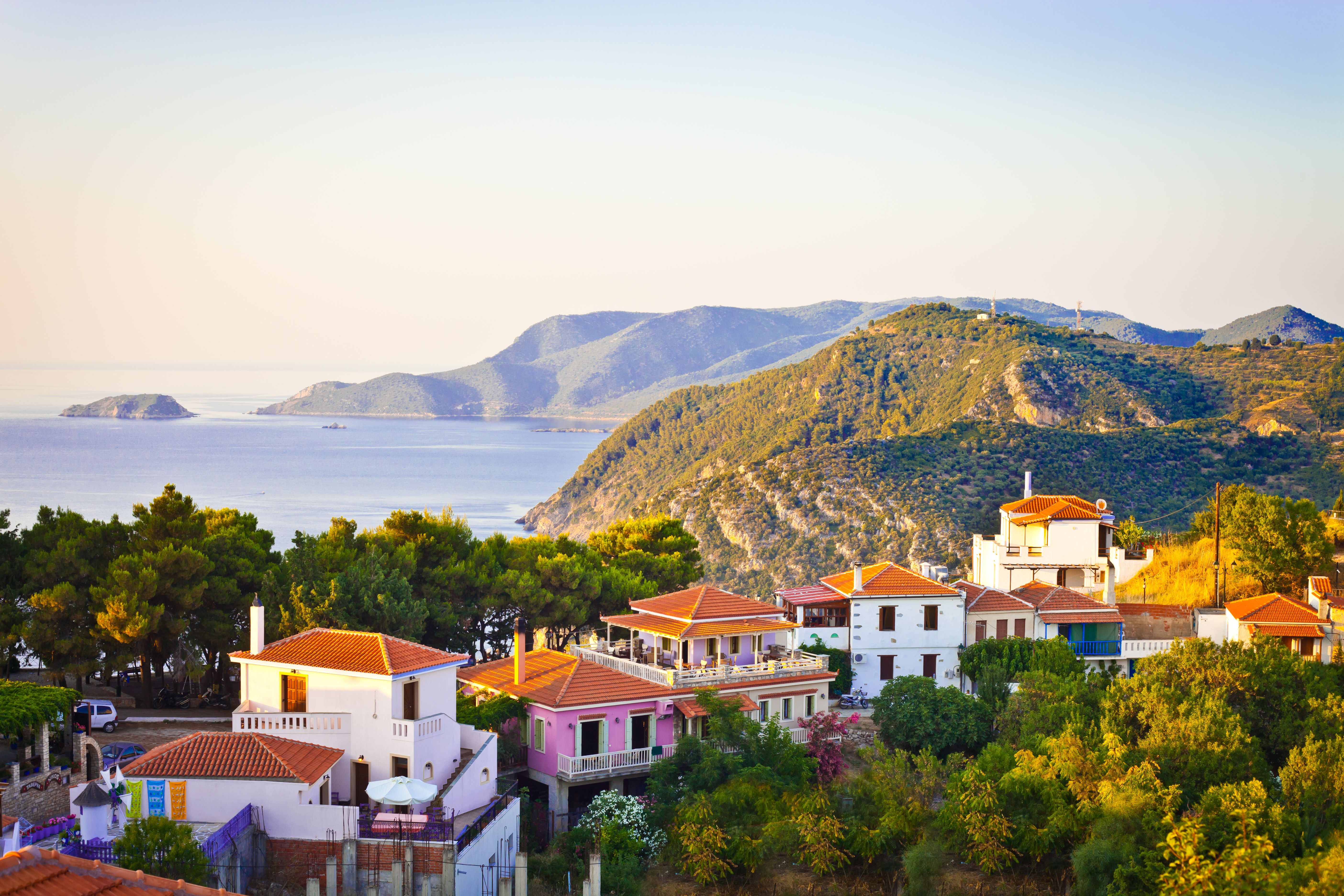 Sunset view of a row of houses tucked between lush trees with grassy hills and a calm sea in the background.