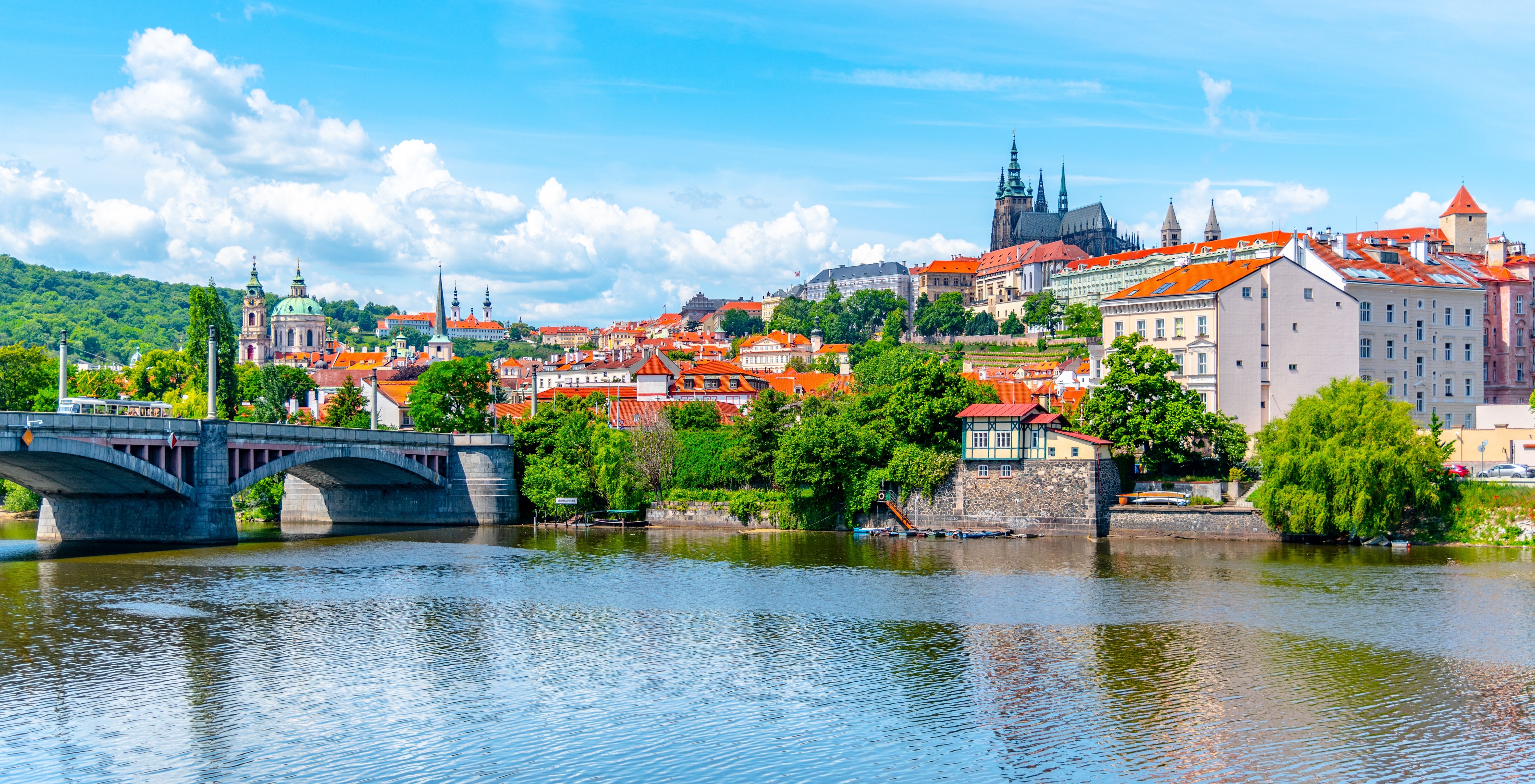 A view of the historic Prague Castle and Manes Bridge on a sunny day with clear skies, overlooking the calm Vltava River in Prague