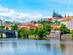 A view of the historic Prague Castle and Manes Bridge on a sunny day with clear skies, overlooking the calm Vltava River in Prague