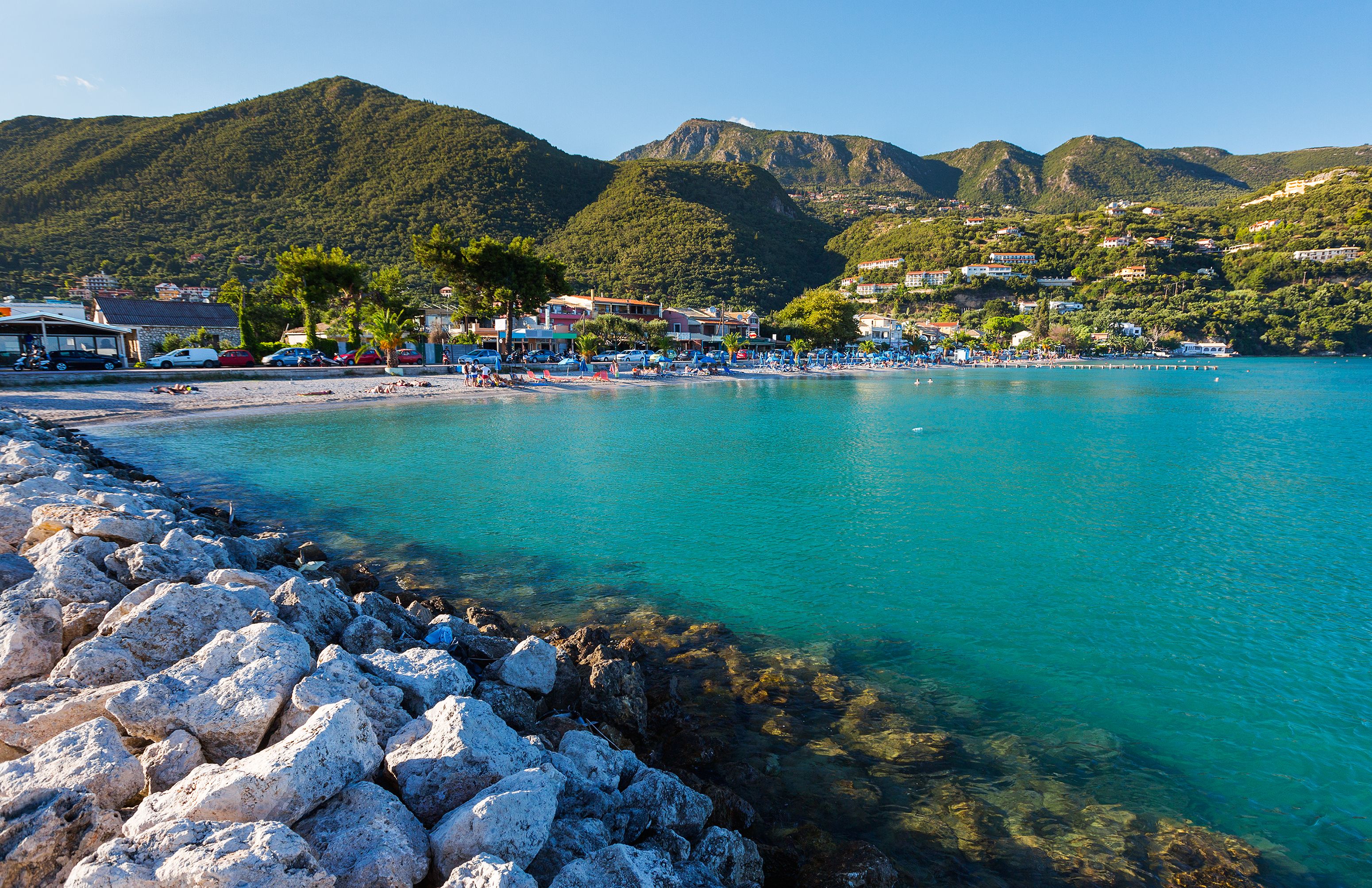 View across calm turquoise water toward a sun-lounger lined beach hemmed in by tree-covered mountains