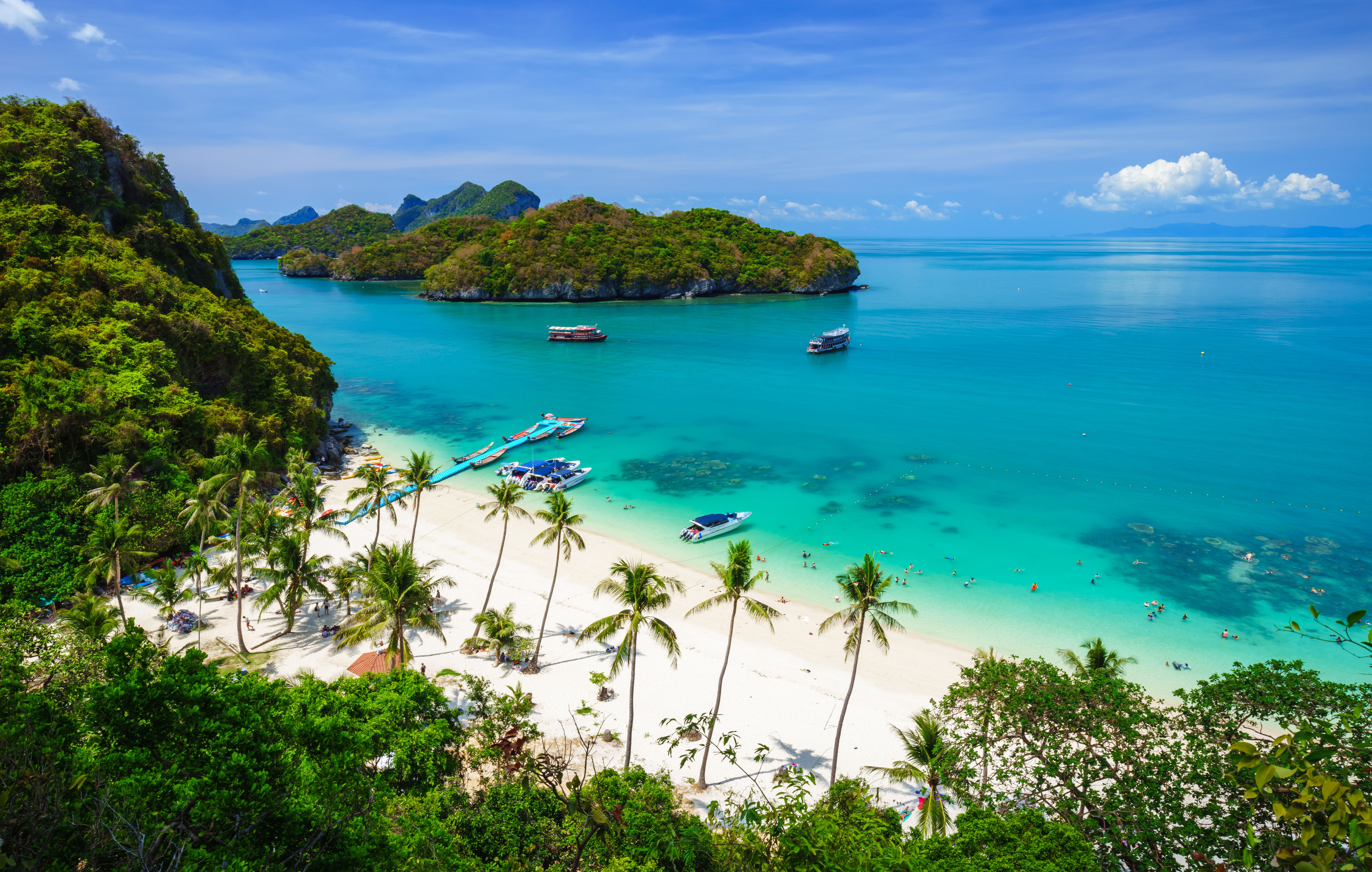 Palm trees, pristine golden sand and clear water on a tropical beach in Thailand with boats and llush green islands in the background
