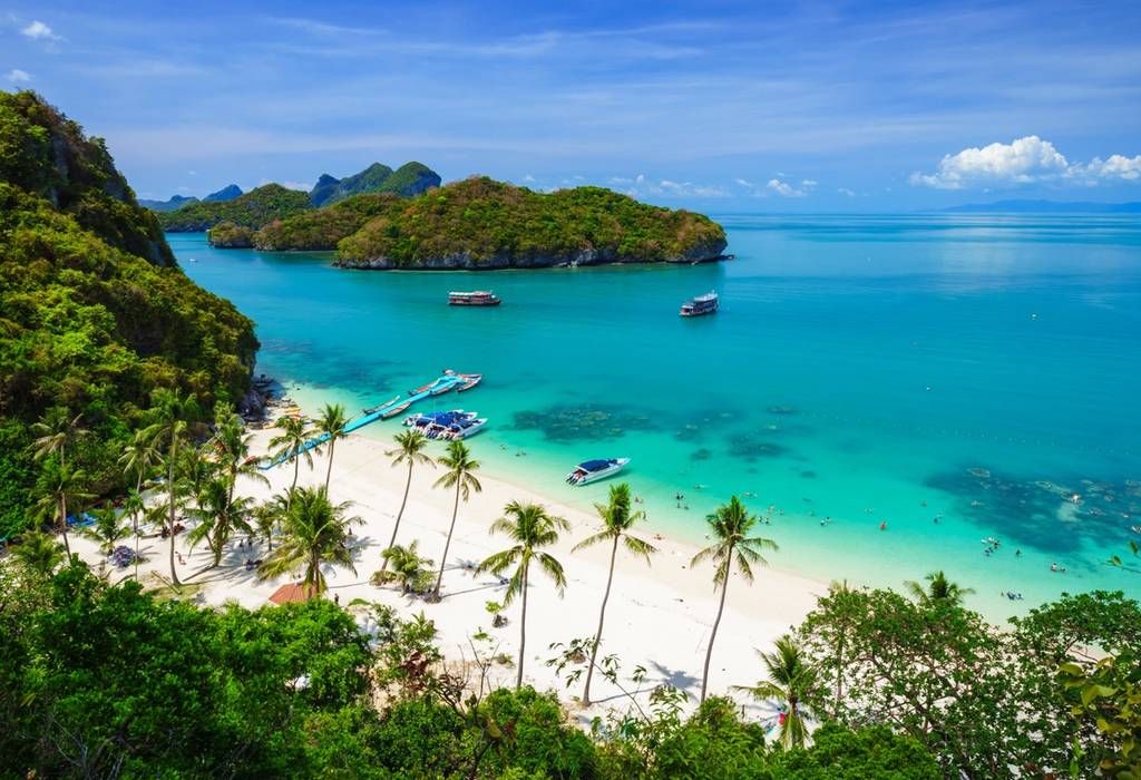 Palm trees, pristine golden sand and clear water on a tropical beach in Thailand with boats and llush green islands in the background