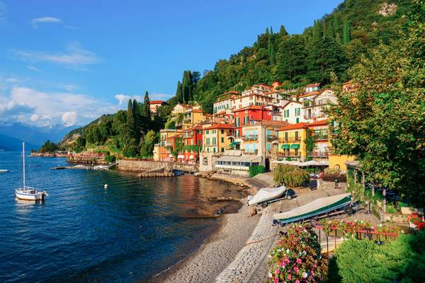 View of a small town perched on the shores of Lake Como with green trees surrounding it and boats bobbing in the water