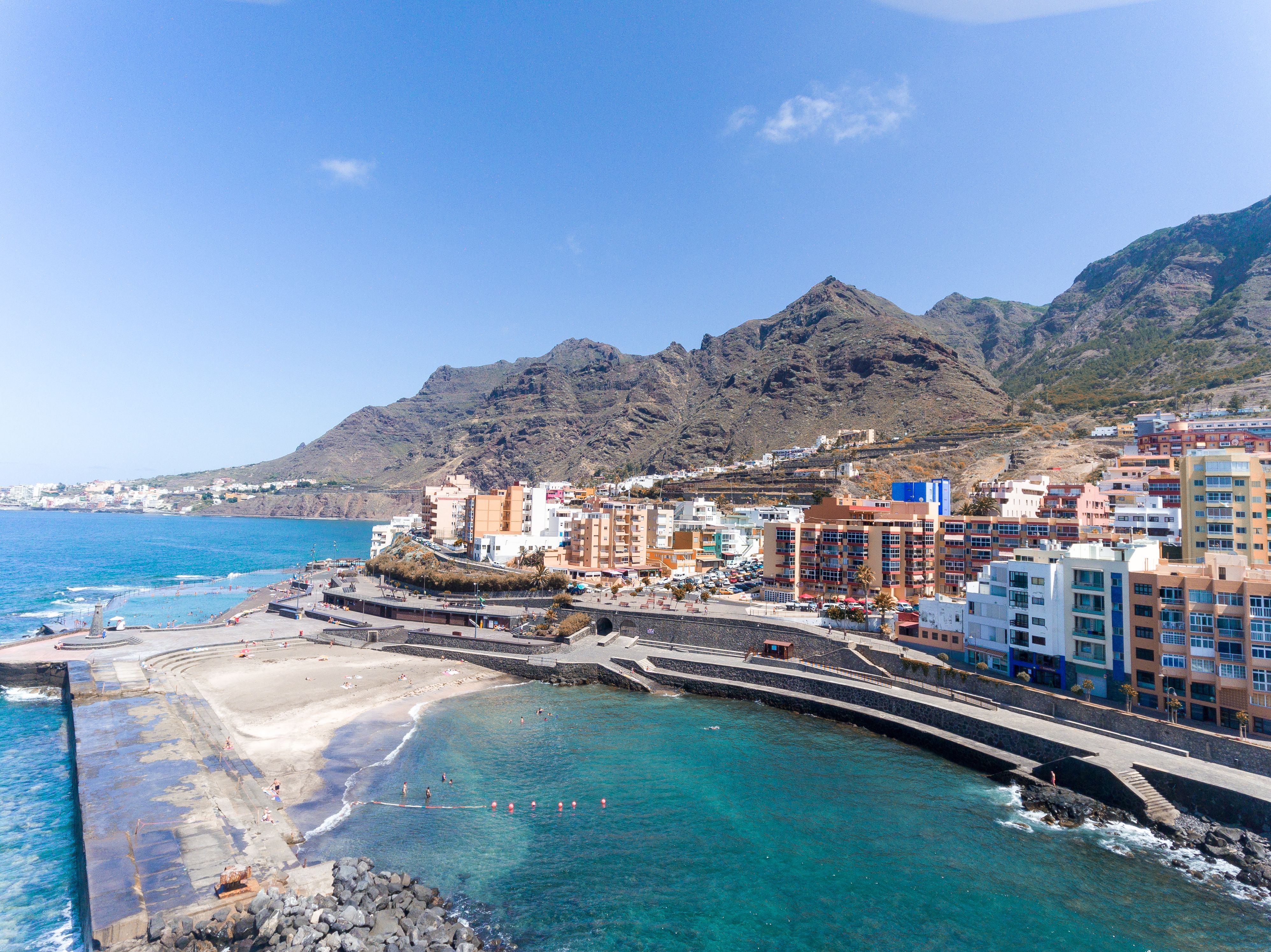 Aerial view of a colourful coastal town fronted by manmade seawater pools and backed by rugged mountains.