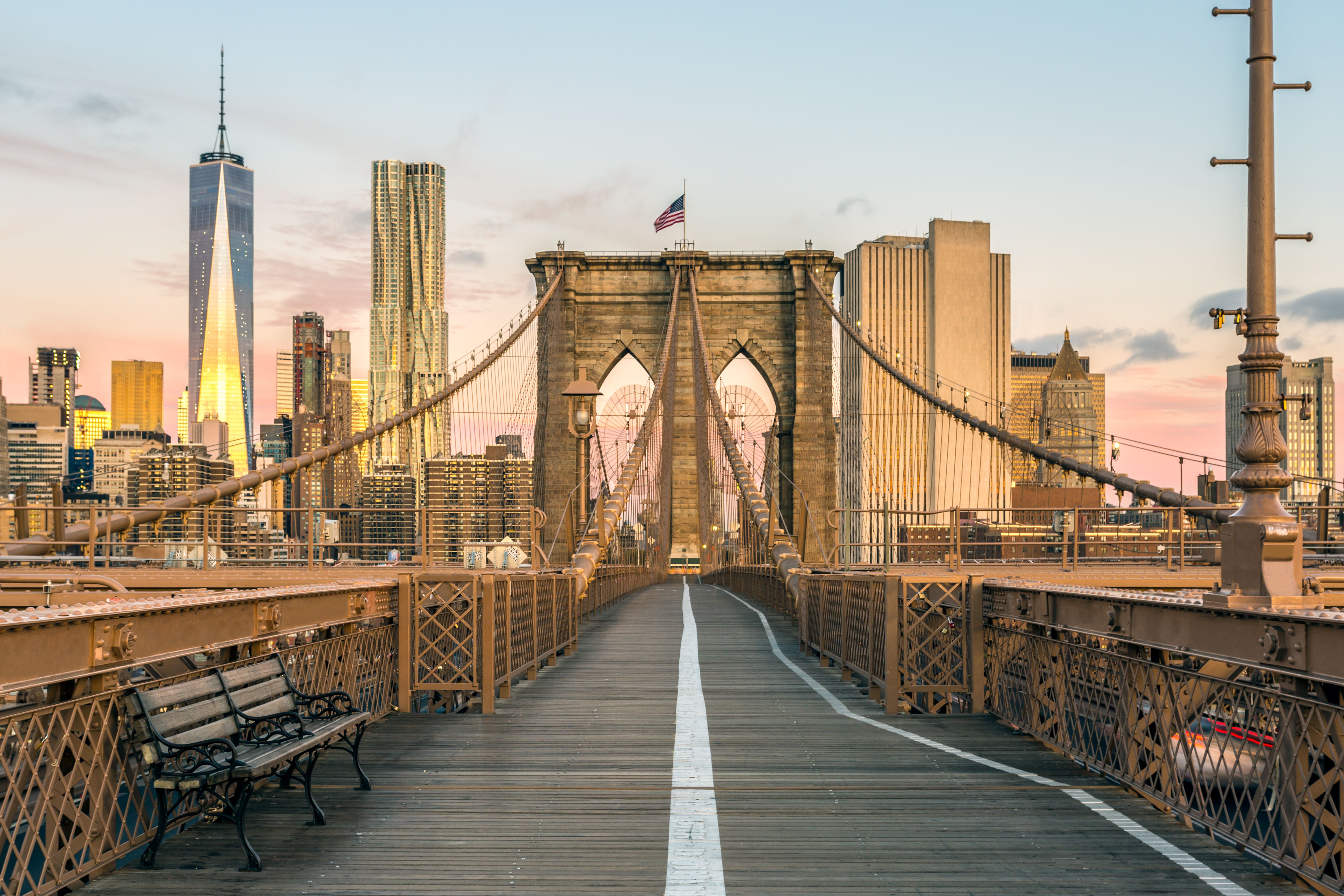 A view looking along the walkway of Brooklyn Bridge in New York as the sun rises over the city's skyscrapers