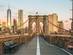 A view looking along the walkway of Brooklyn Bridge in New York as the sun rises over the city's skyscrapers