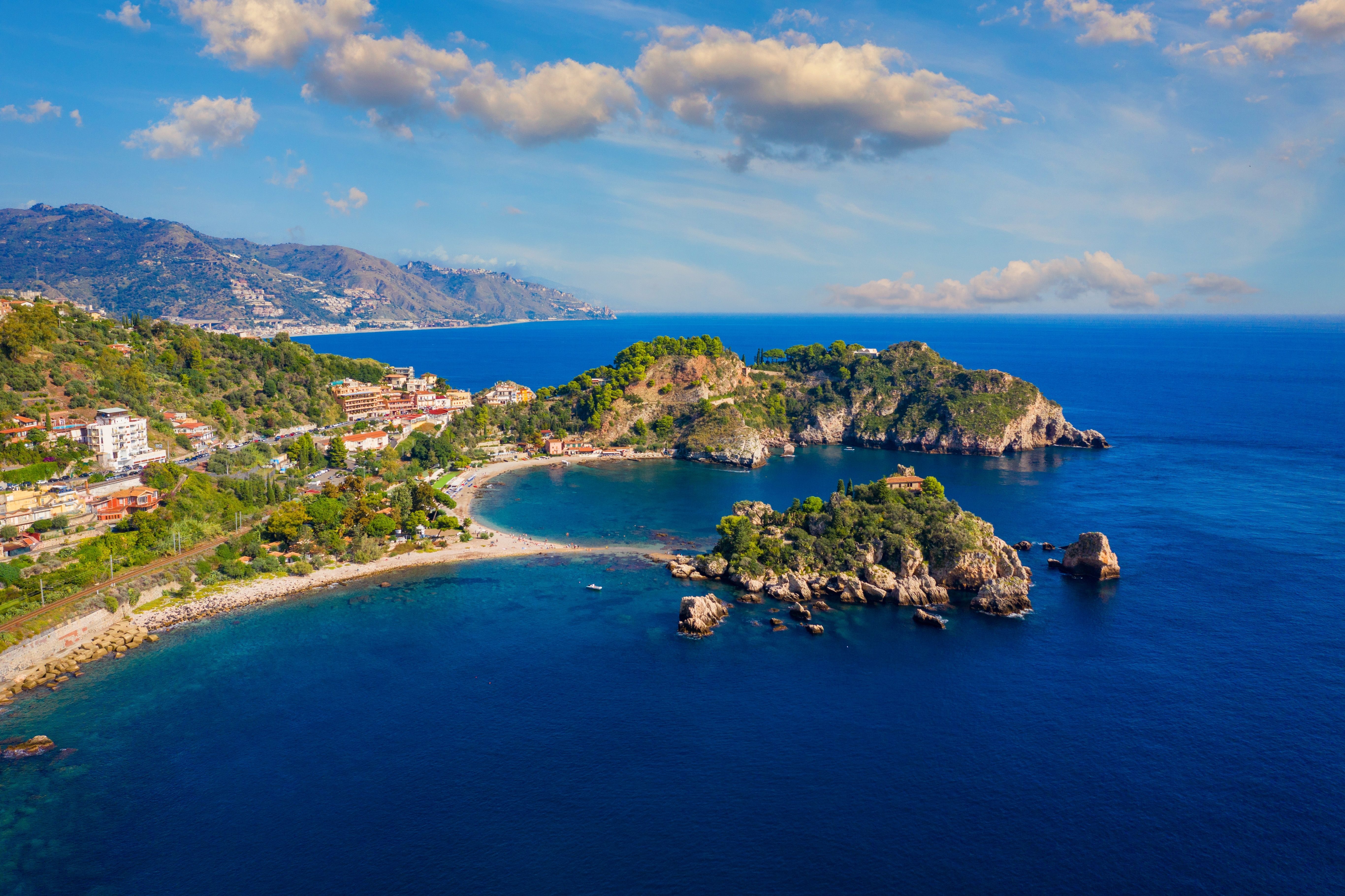 A narrow path connects a rocky island to a mainland with two curved beaches.