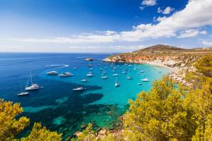 A picturesque view of Cala d Hort beach in Balearic Islands, Ibiza, Spain with many anchored yachts in clear blue water on a sunny day
