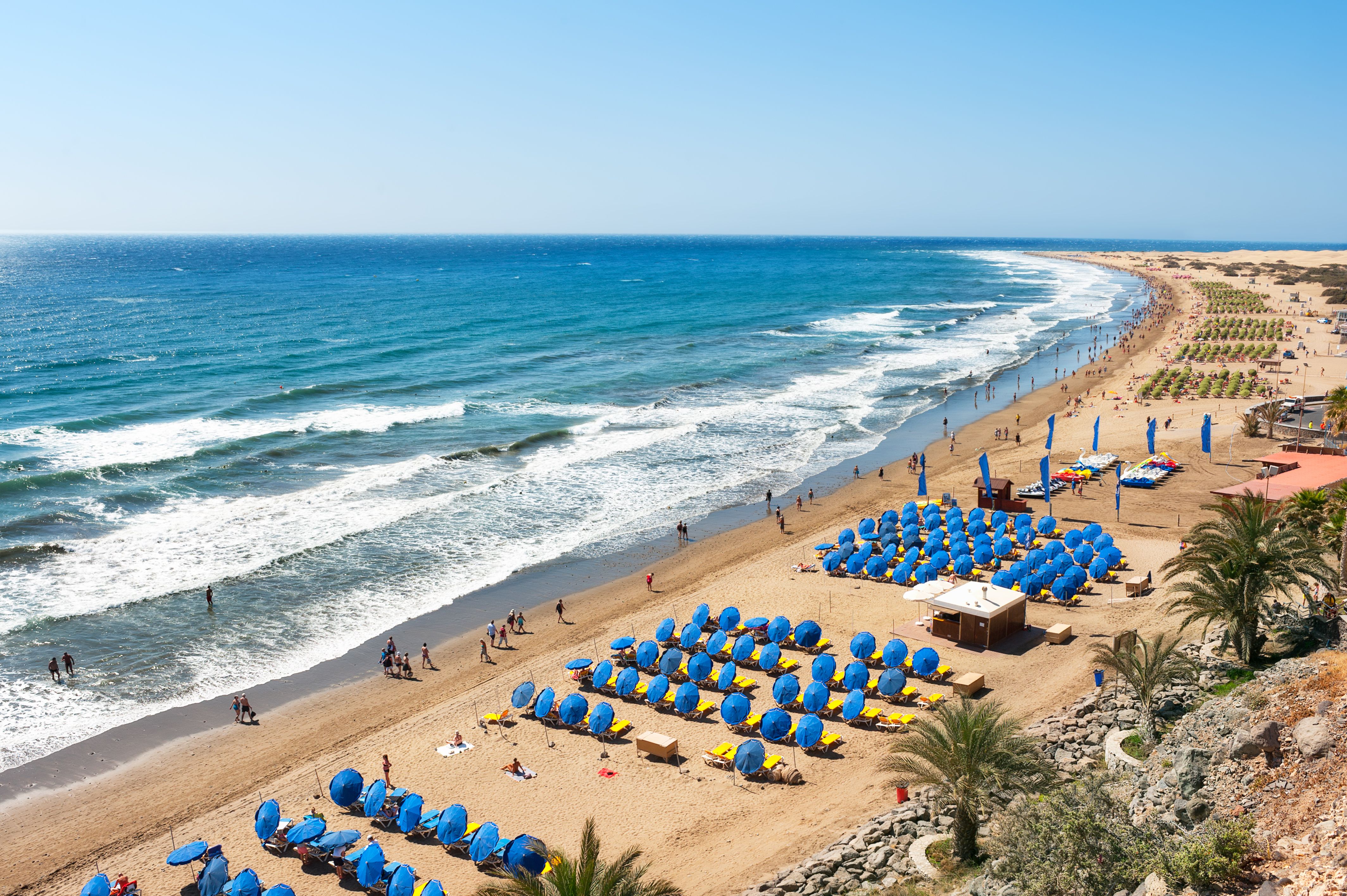 A view of Playa del Ingles in Gran Canaria with blue umbrellas and sunbeams lining the beach on a clear blue day