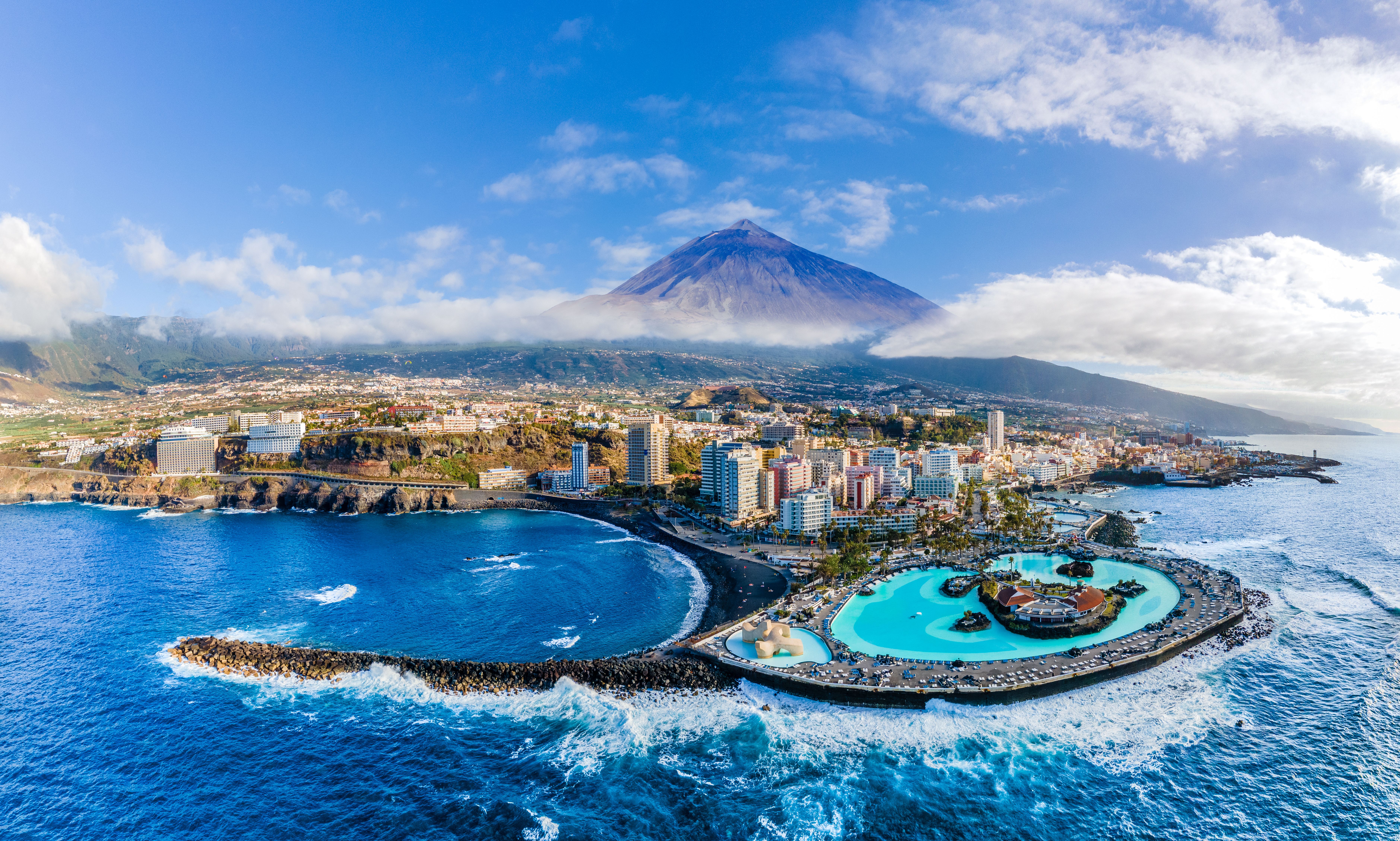 View of Mount Teide bearing over the city of Puerto de le Cruz in Tenerife, complete with a curved black-sand beach and the artifical saltwater pools of the César Manrique Maritime Park 