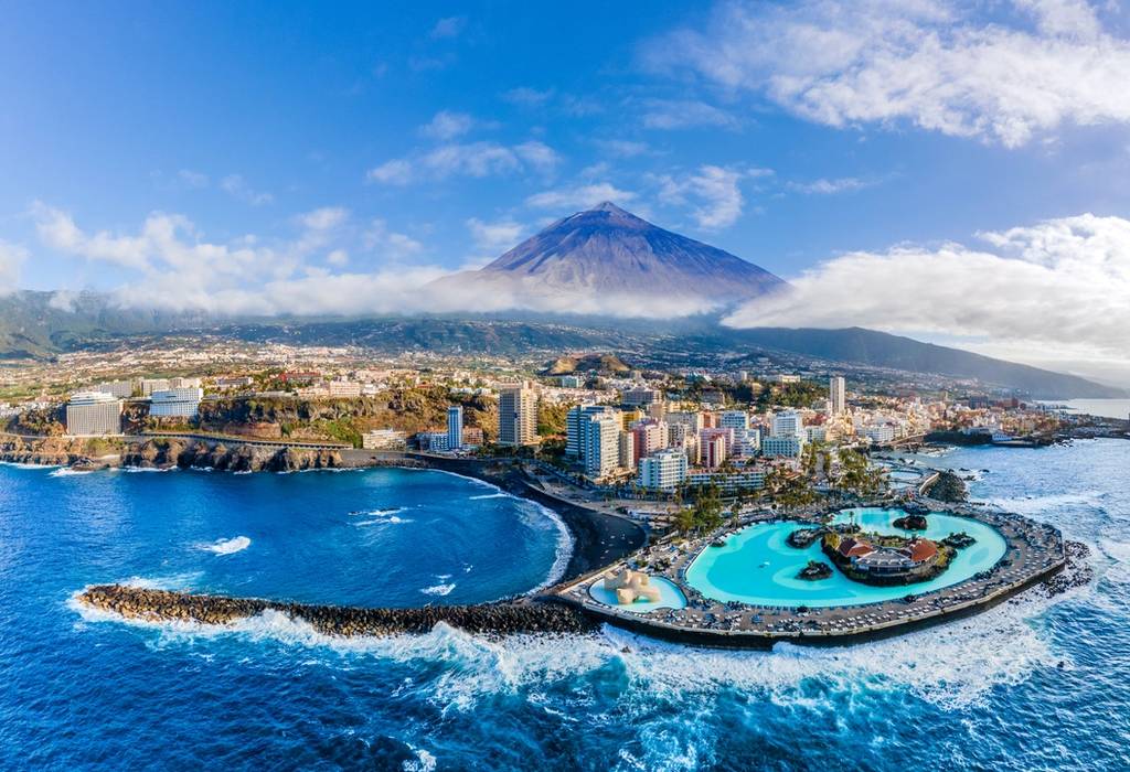 View of Mount Teide bearing over the city of Puerto de le Cruz in Tenerife, complete with a curved black-sand beach and the artifical saltwater pools of the César Manrique Maritime Park 