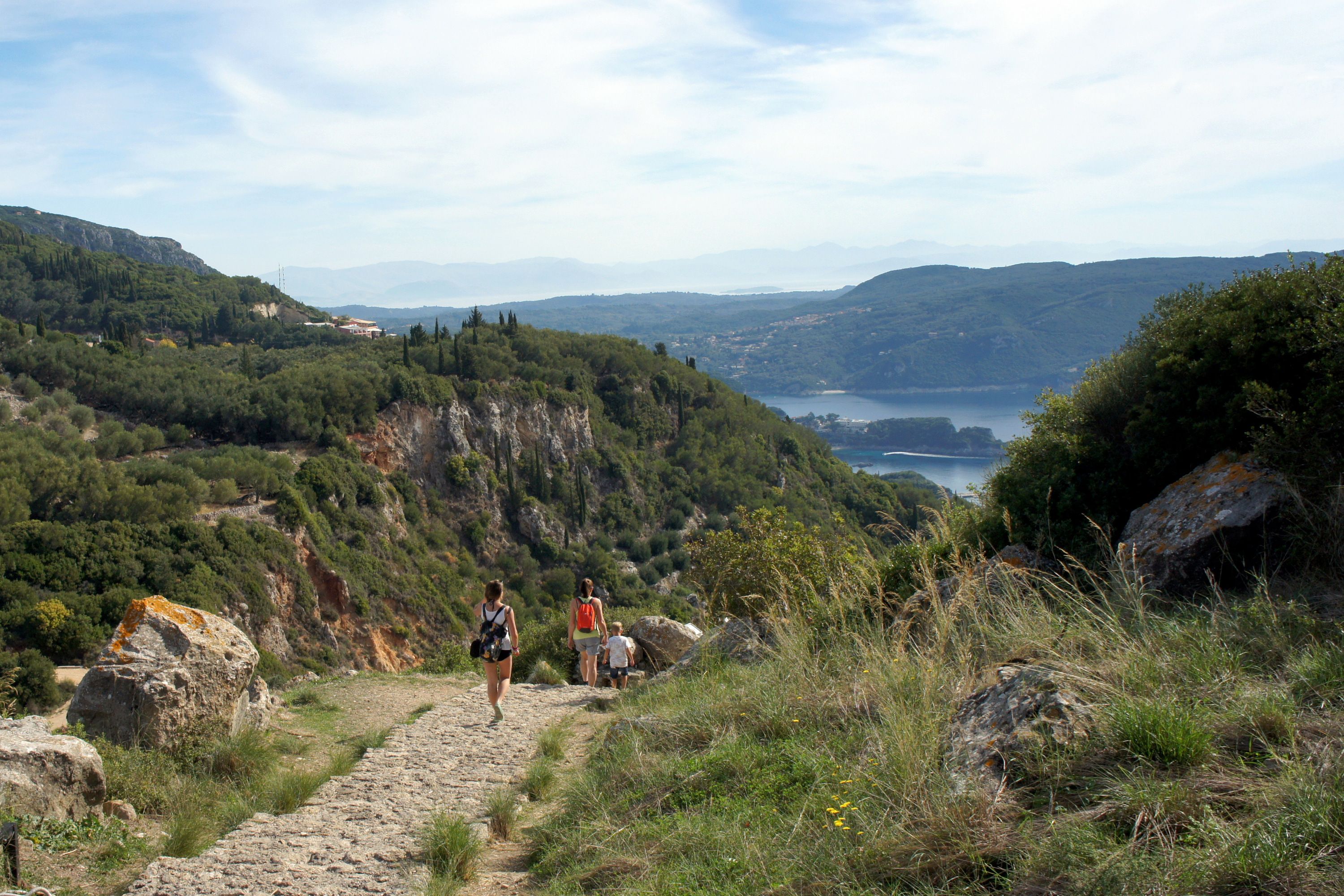 Two women and a child hiking along a greenery-surrounded trail towards the coast beyond