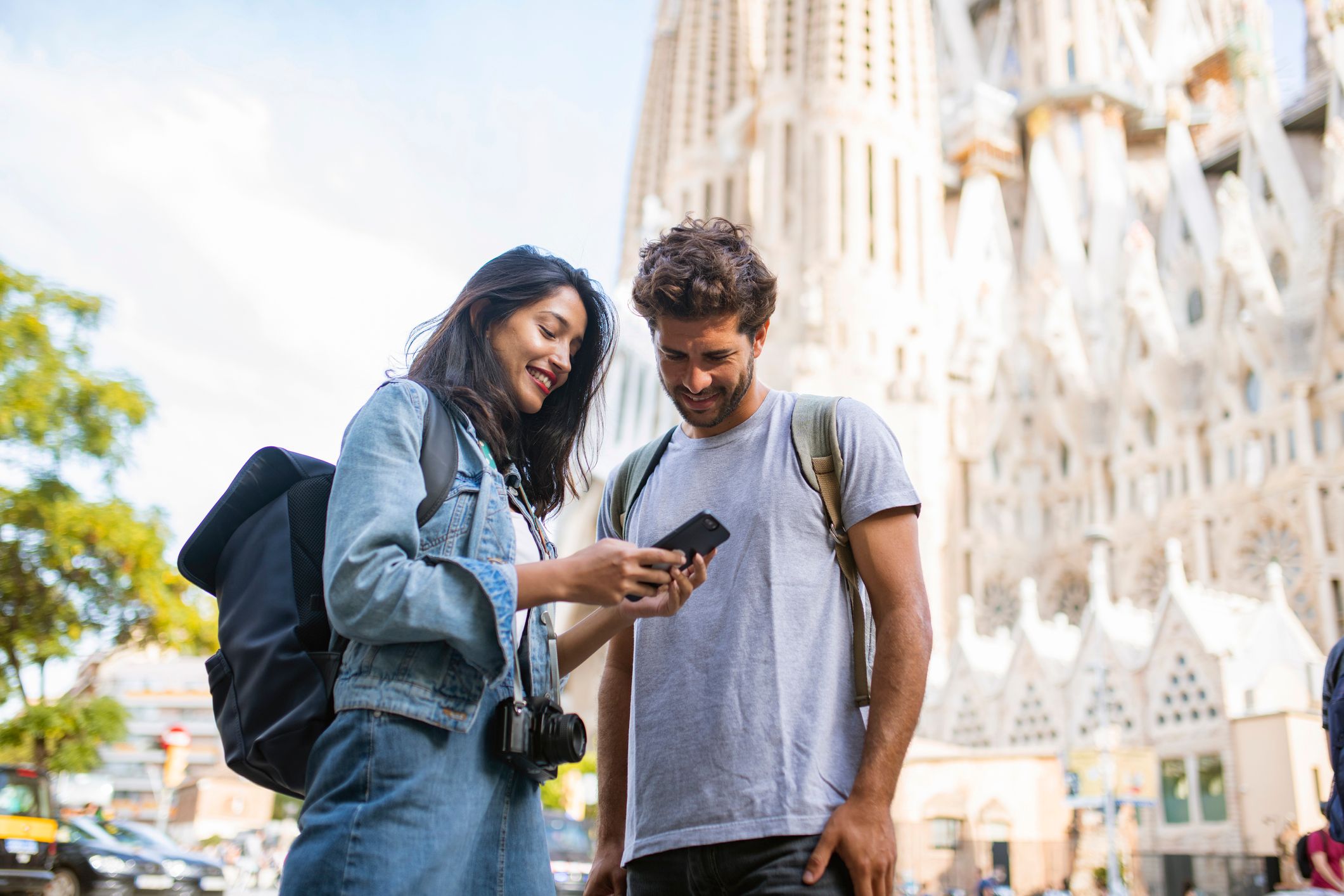 A picture of a couple sightseeing in Barcelona and smiling as they look at smartphone with La Sagrada Familia in the background