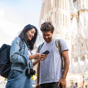 A picture of a couple sightseeing in Barcelona and smiling as they look at smartphone with La Sagrada Familia in the background