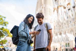 A picture of a couple sightseeing in Barcelona and smiling as they look at smartphone with La Sagrada Familia in the background