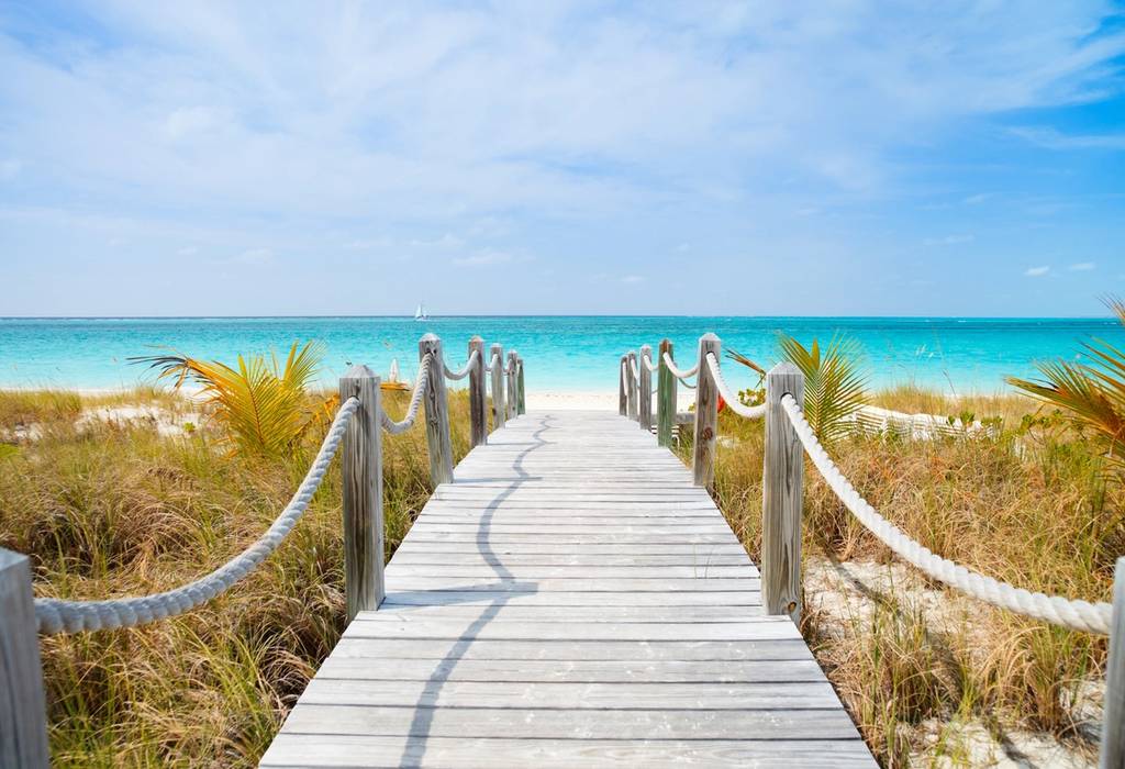A boardwalk leading between grasses and across sand to a white-sand beach with bright blue waters in the Caribbean