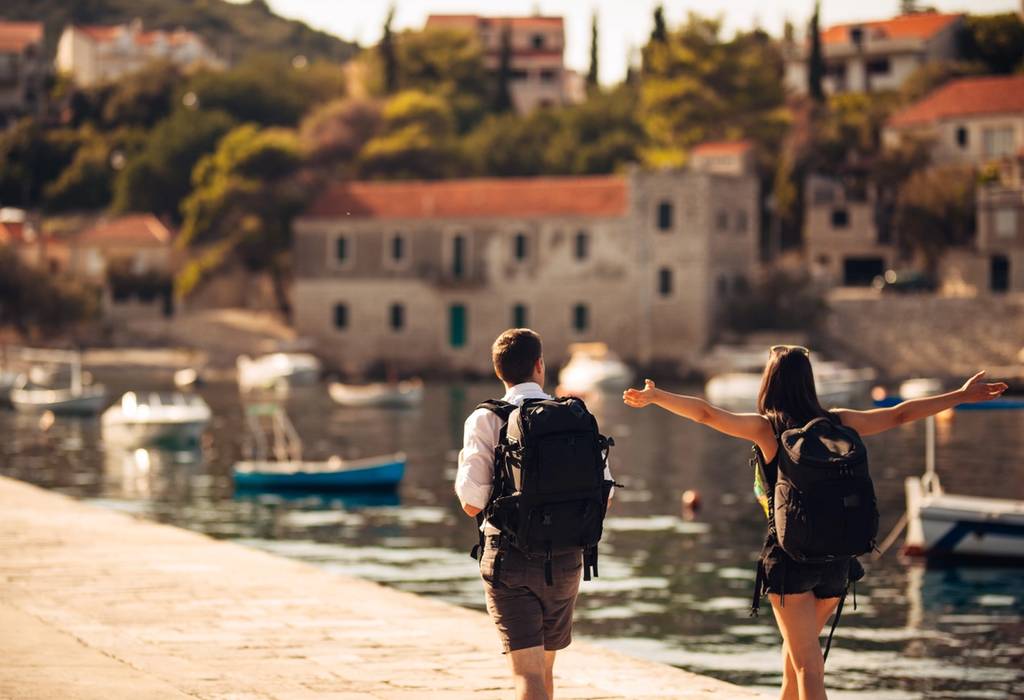 Two young backpackers walking along a tiny harbour filled with fishing boats with traditional stone houses in the background