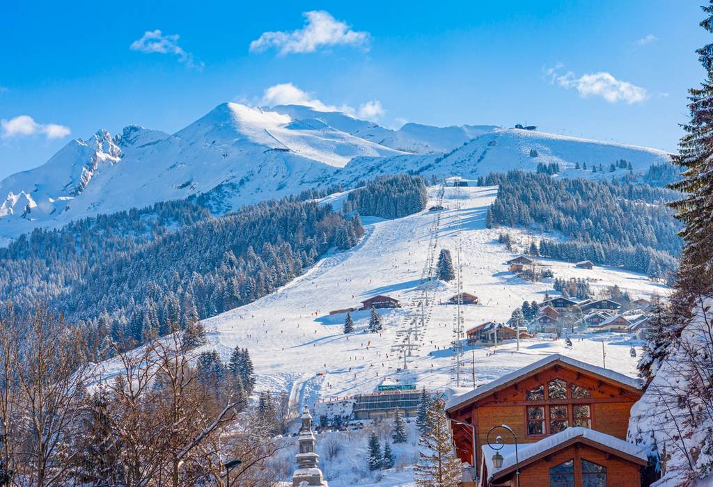 A view of La Clusaz Ski slopes on a sunny winter day in the French Alps with a telecabin gondola, people skiing and chalets in the foreground with mountains and ski slopes covered in snow