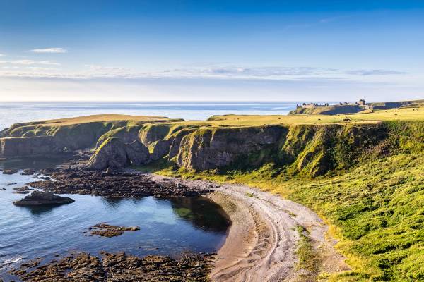 View of the green cliffs and small beach at Strathlethan Bay with Dunnottar Castle in the distance