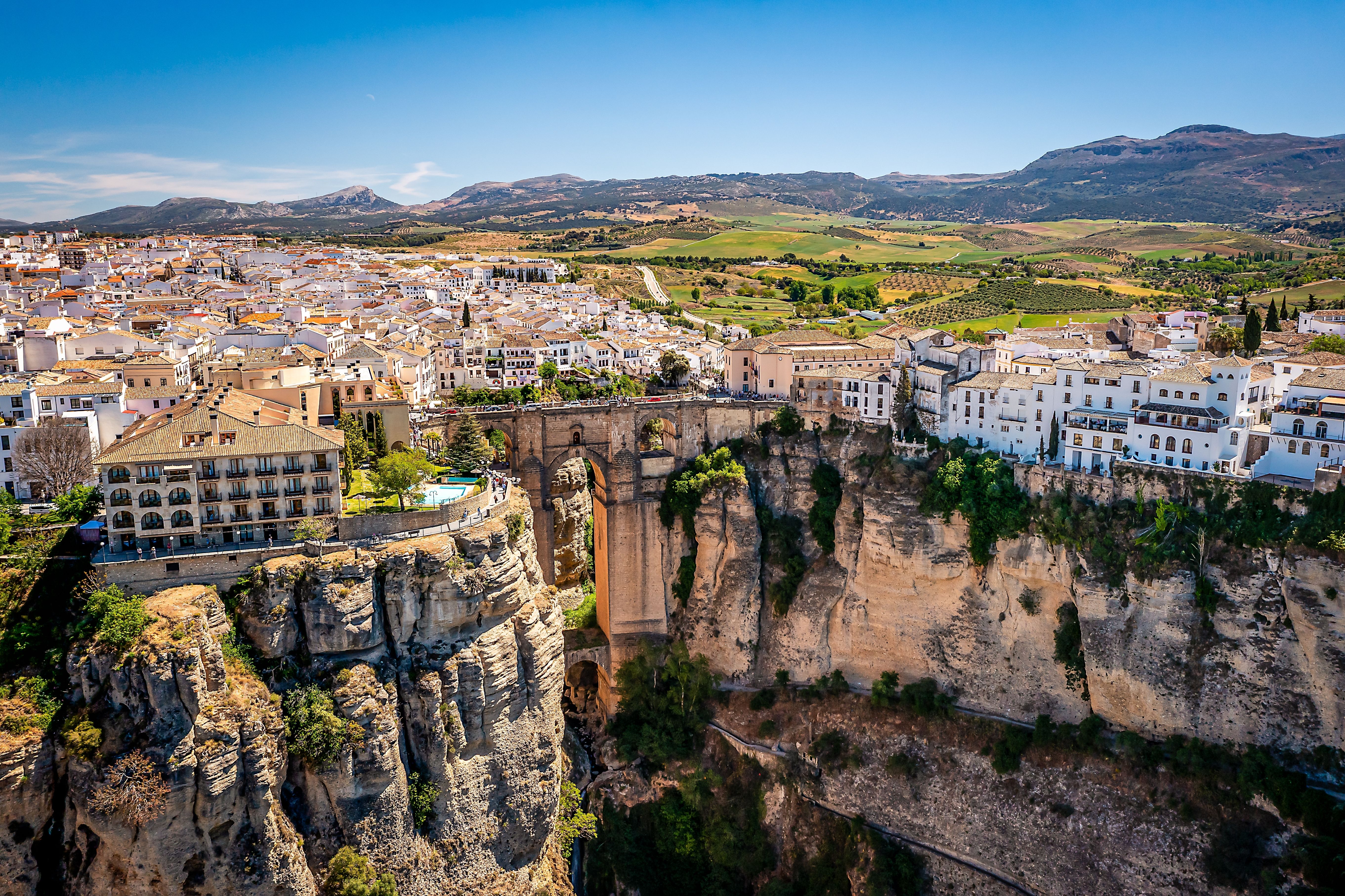 Aerial view of a whitewashed Spanish village perched on a huge canyon and connected by a giant bridge.
