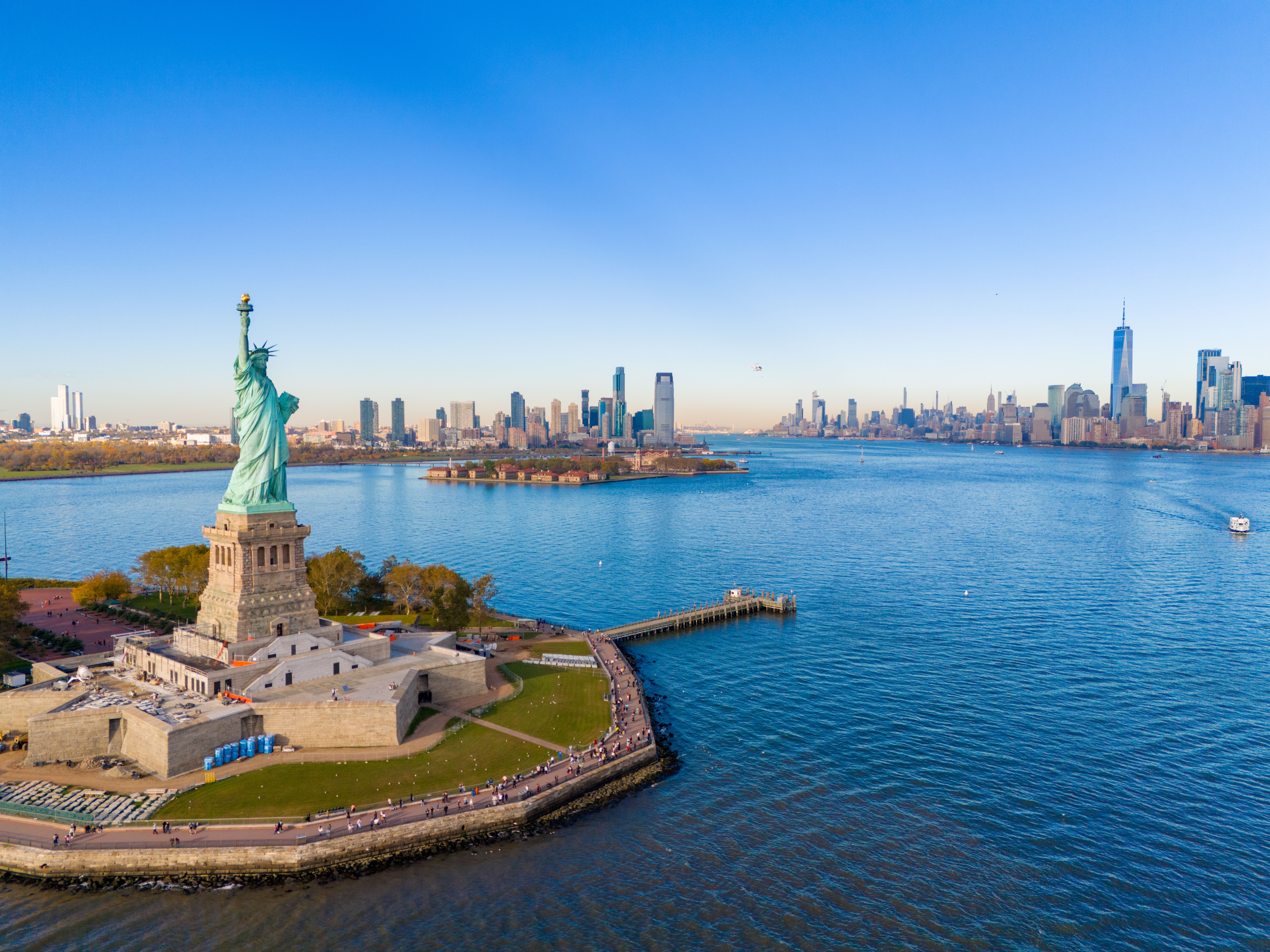 Aerial view of the Hudson Bay with the Statue of Liberty in the foreground and New York City's skyscrapers beyond.