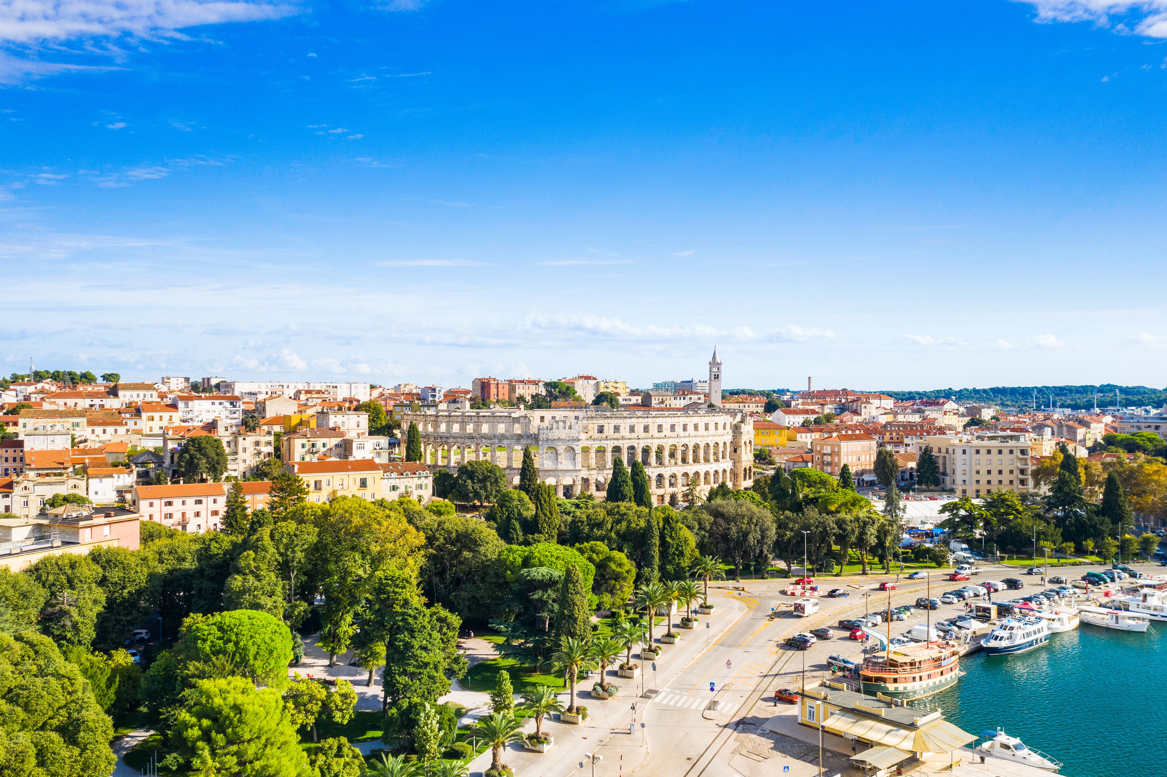 A panoramic view of an ancient Roman amphitheatre, the old town centre of Pula and a small boat-lined harbour on a sunny day